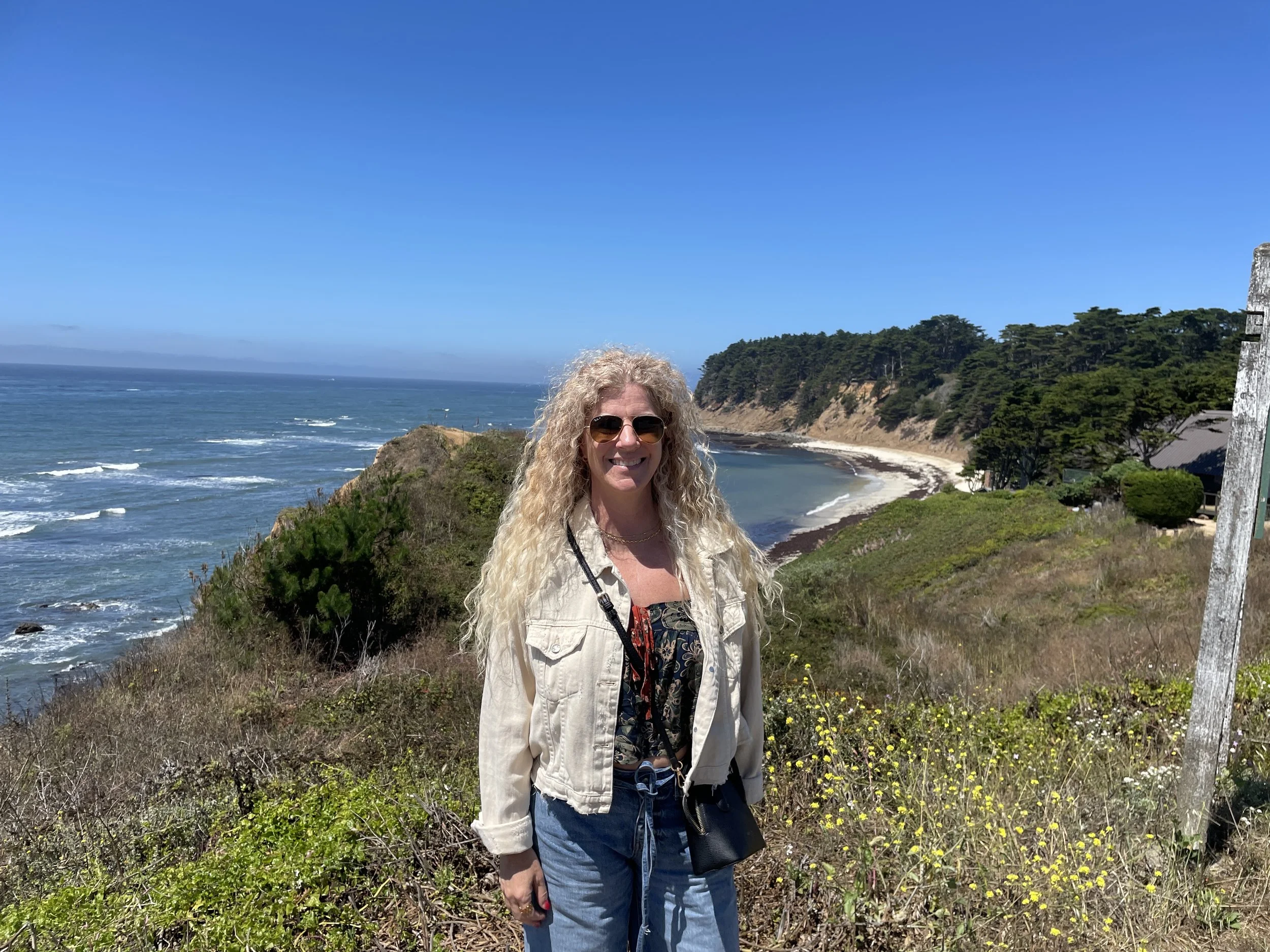Woman with curly blonde hair wearing sunglasses, a beige jacket, and blue jeans standing near cliffs and ocean on a sunny day at a coastal viewpoint.