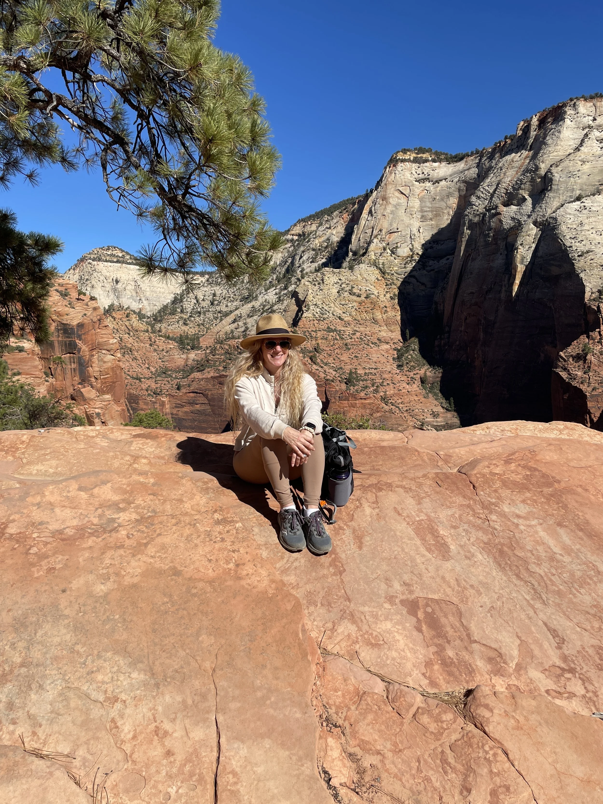 A woman wearing a hat, sunglasses, and hiking clothes, sitting on a large, smooth, red rock in a desert canyon landscape with cliffs, sparse trees, and a clear blue sky.