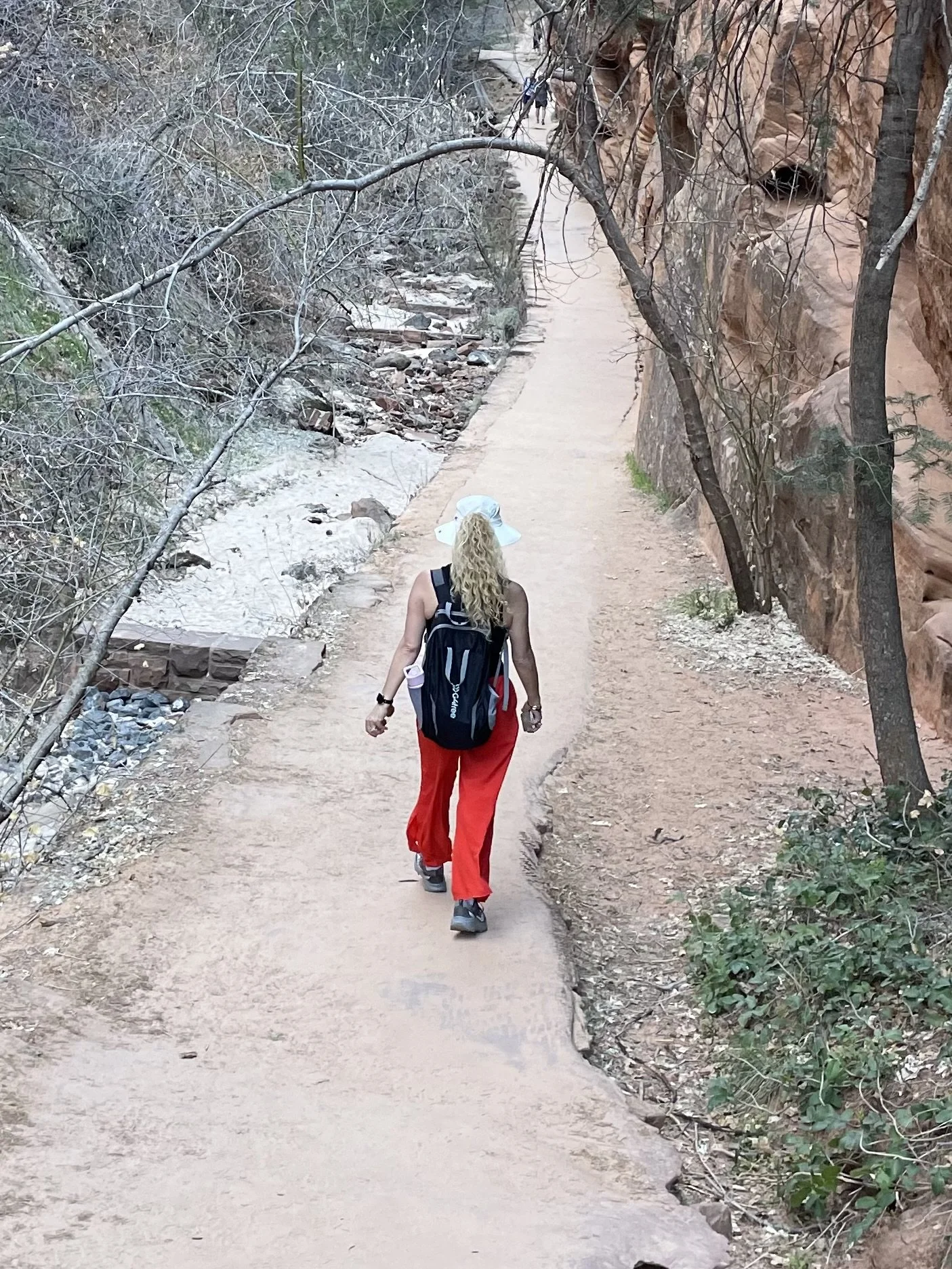 A woman with curly blonde hair wearing a large white sun hat, black backpack, and red pants walking along a sandy trail in a red rock canyon with sparse vegetation and rock formations on the right side.