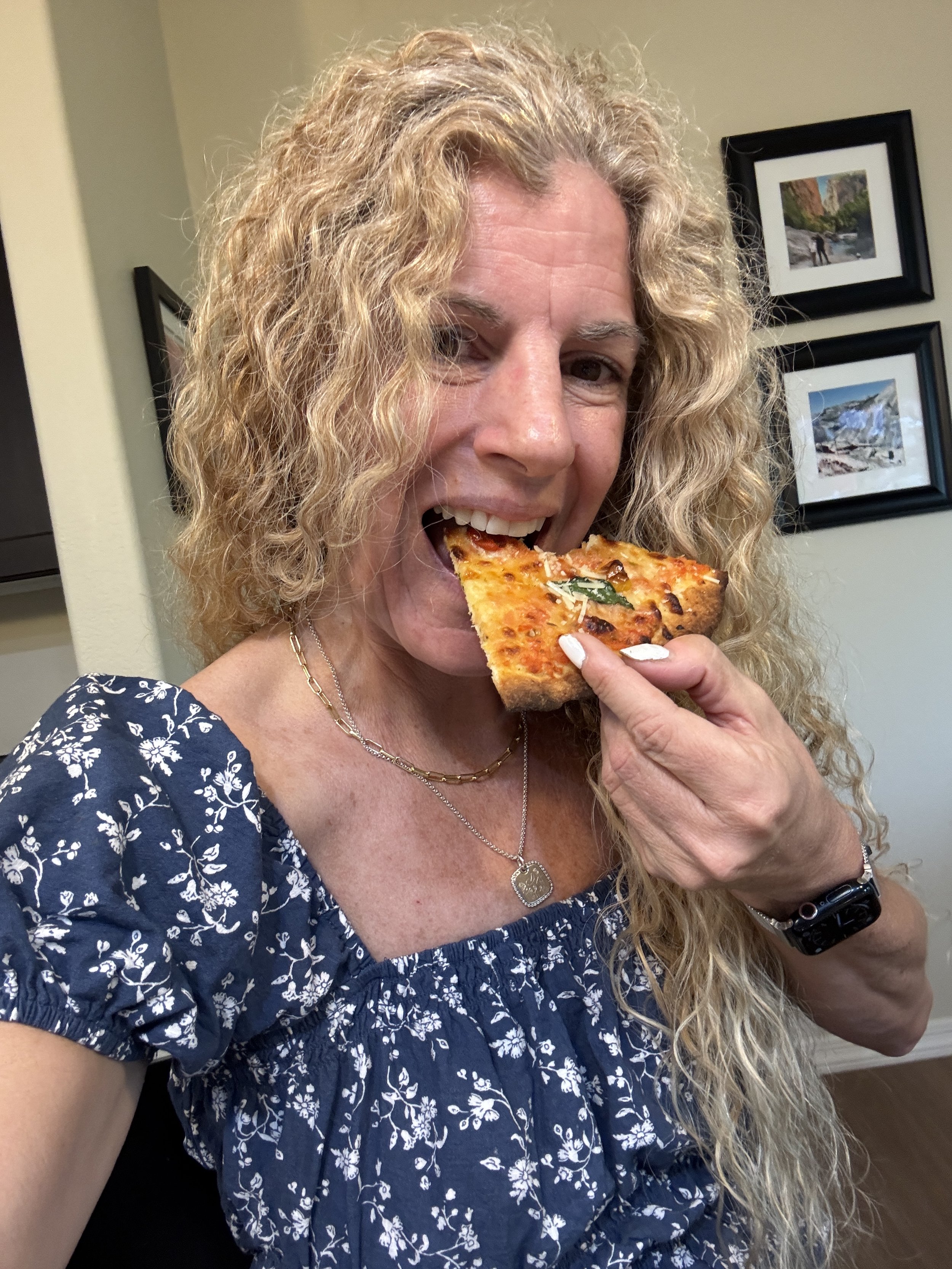 A woman with long, curly blonde hair wearing a navy blue dress with white floral print, holding a slice of pizza near her mouth, smiling, indoors with framed pictures on the wall.