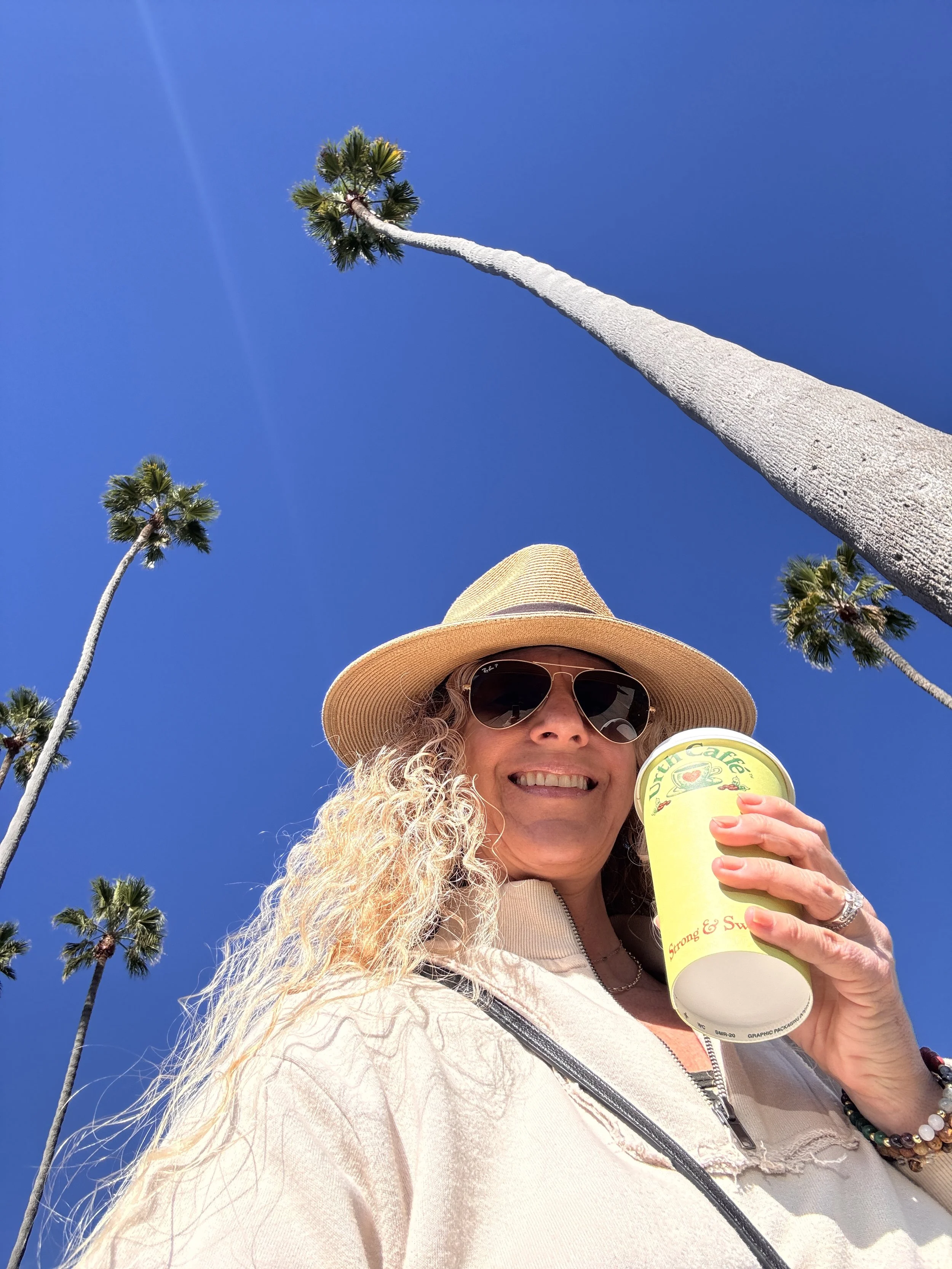 A woman with curly blonde hair wearing a wide-brimmed straw hat, sunglasses, and a white jacket, holding a yellow cup, smiling, with palm trees against a clear blue sky in the background.