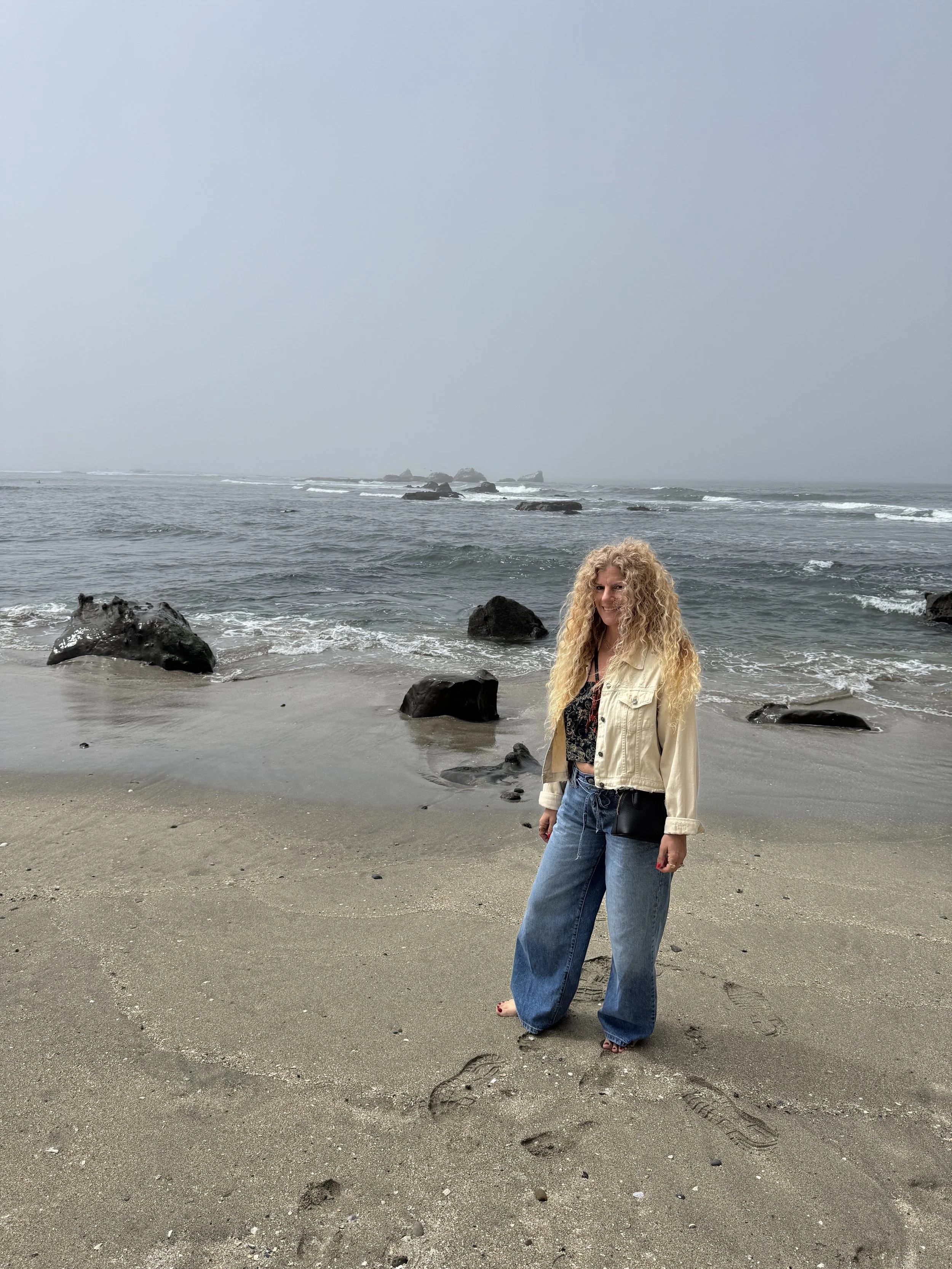 A woman with curly blonde hair standing on a sandy beach near the ocean, wearing a light-colored jacket and loose blue jeans, with ocean waves and rocks in the background.