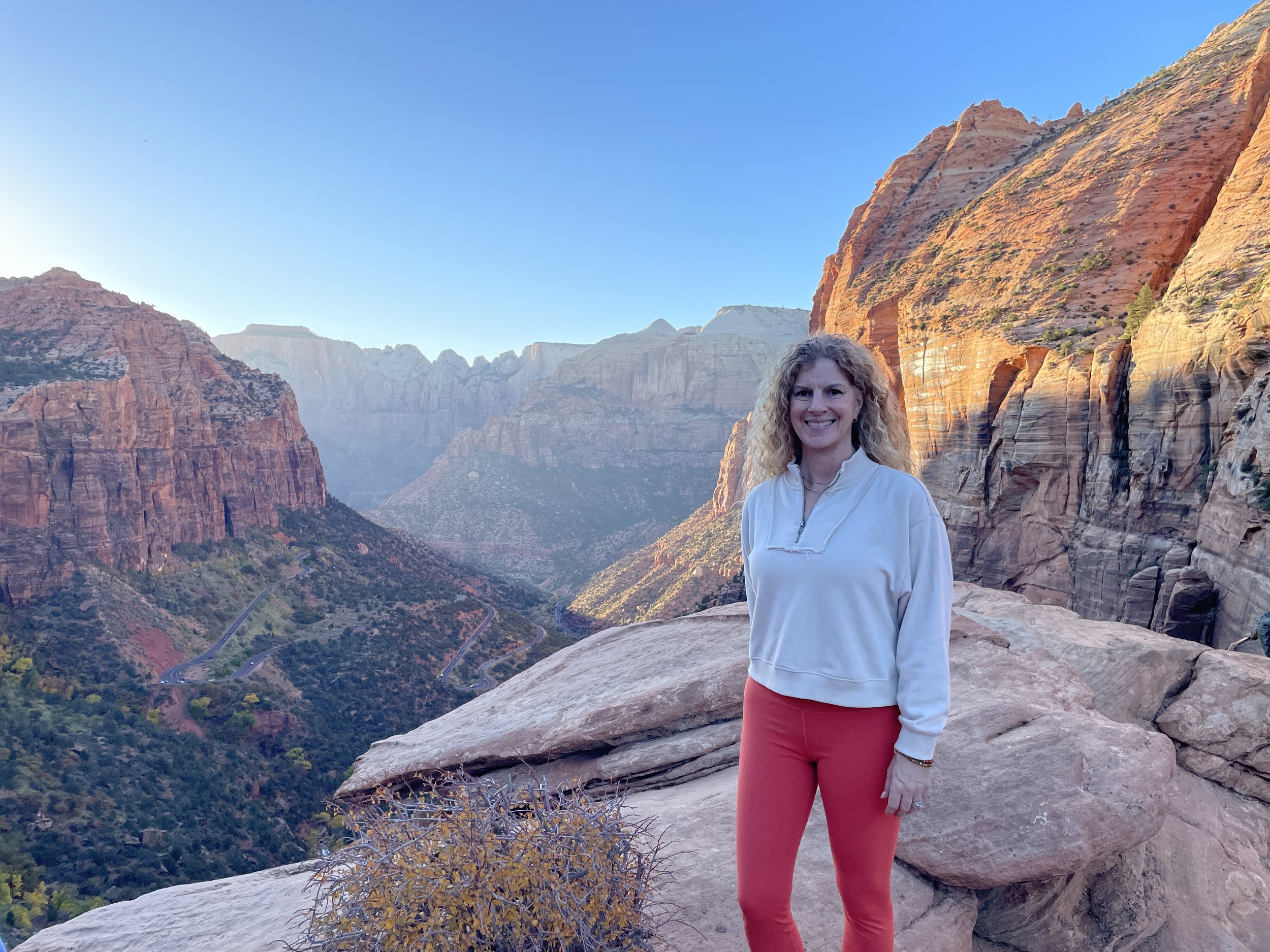 A woman with curly blonde hair smiling at the camera while standing on a rocky ledge in a canyon with red rock formations and a winding road below, under a clear blue sky.