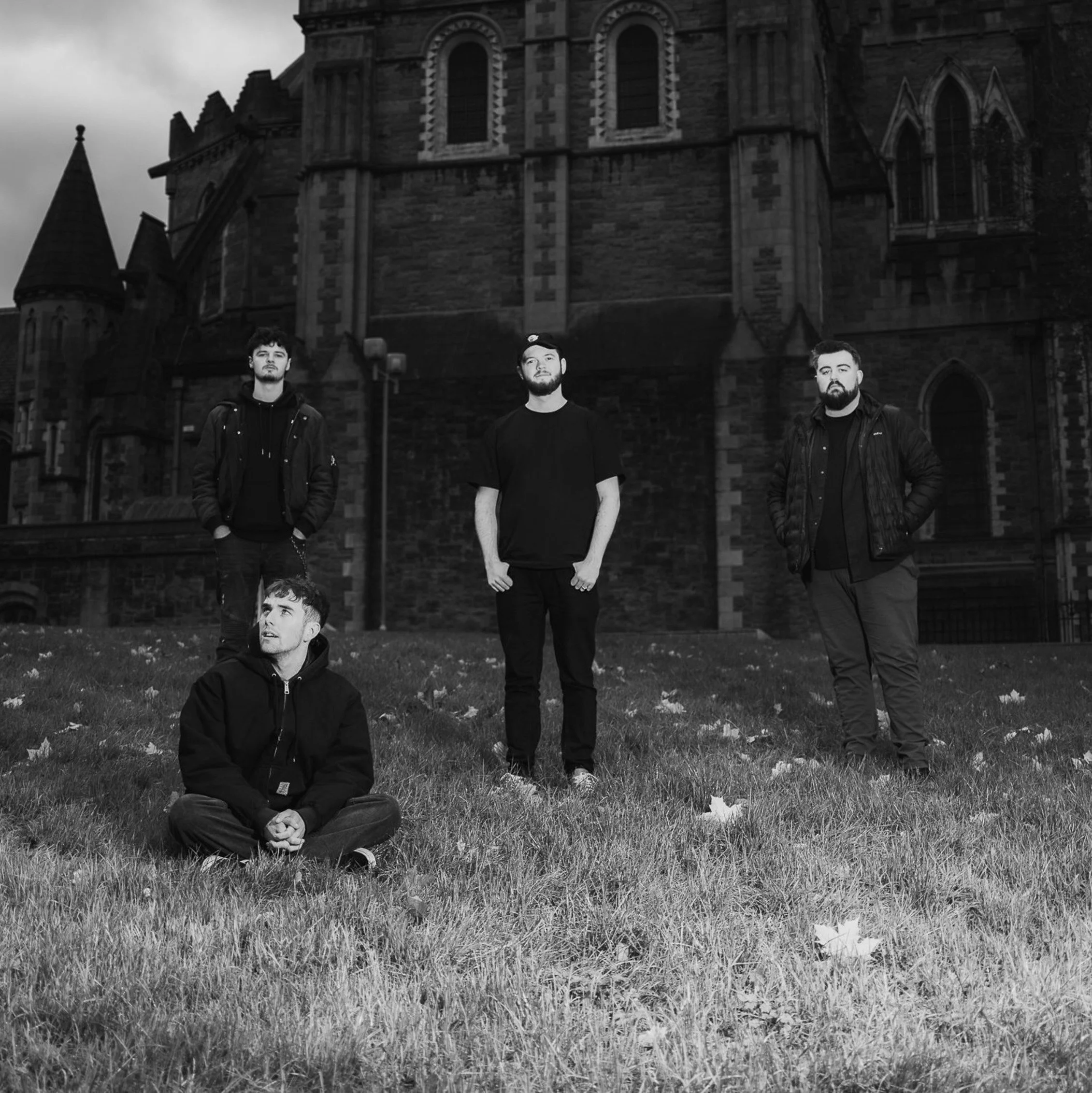 A black and white photograph of three young men standing and sitting on a grassy area in front of an old stone church or castle with gothic architecture, pointed towers, and arched windows during daytime.