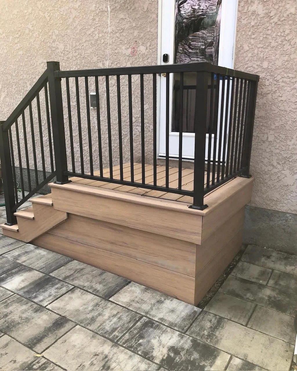 A small porch with a black metal railing, wooden flooring, and steps leading up to a white door.