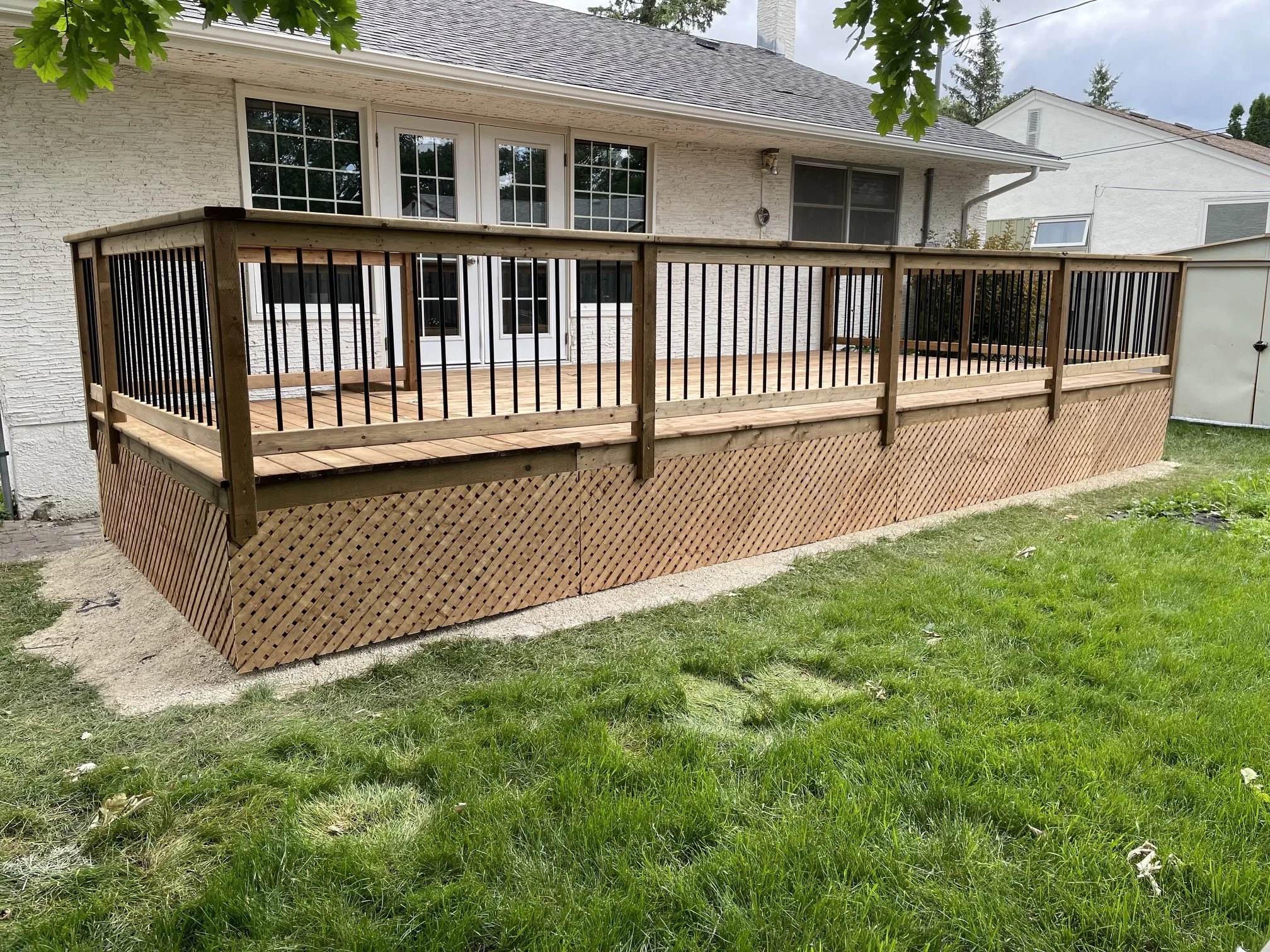 A wooden deck attached to a white brick house with a sliding glass door, overlooking a grassy backyard with trees.