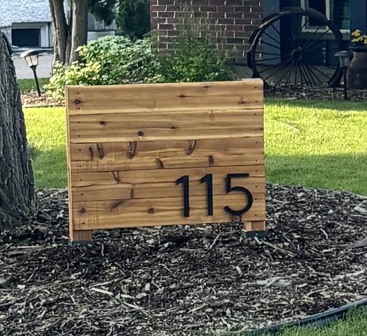 Wooden house number sign displaying 115, placed in a landscaped garden with shrubs, trees, a brick wall, and vintage wheel decoration in the background.