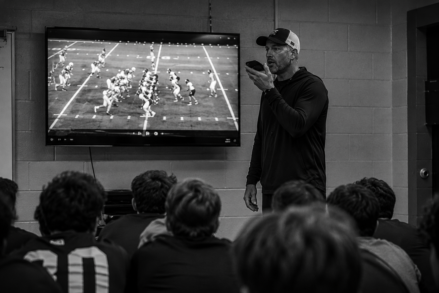 Football coach giving a presentation to a team in a locker room, with a large screen displaying a football game diagram.