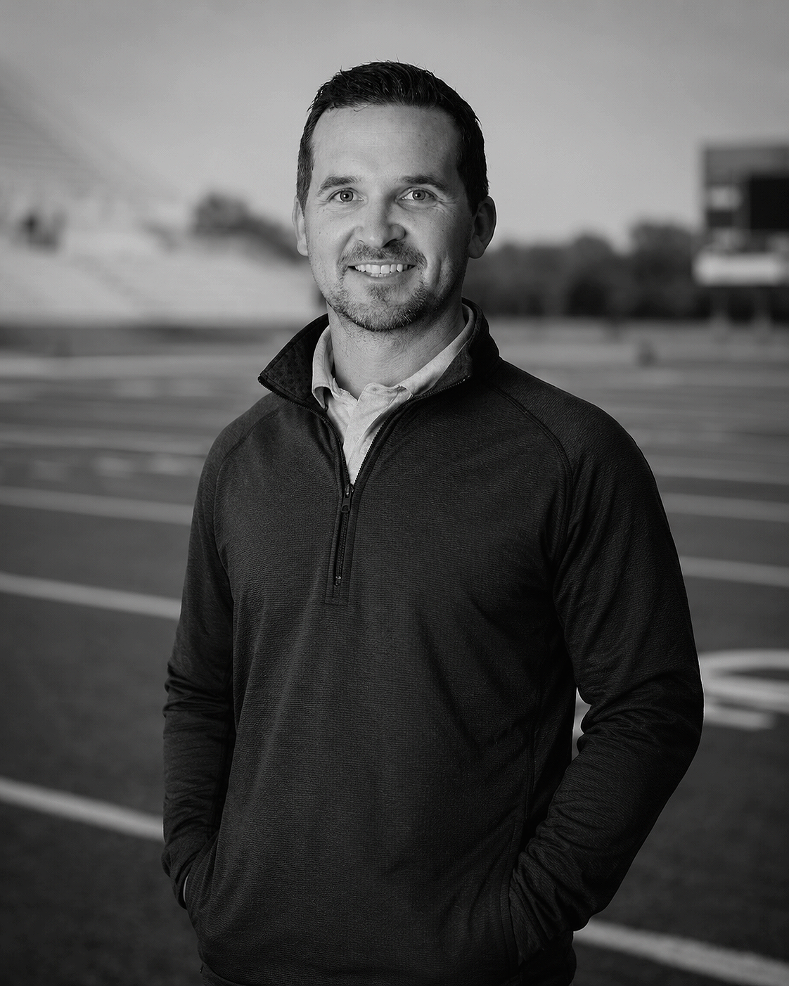 Black and white photo of a man standing on a track field, smiling, with trees and buildings in the background.