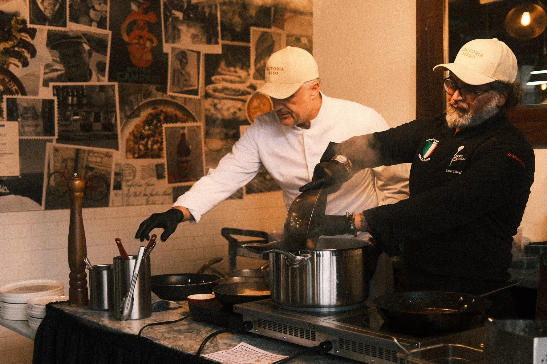 Two men wearing white caps and black gloves cooking at a restaurant kitchen. One man is wearing a white chef's coat, and the other is wearing a black jacket with logos. They are working with pots on a stove, with steam rising from the food. The backg