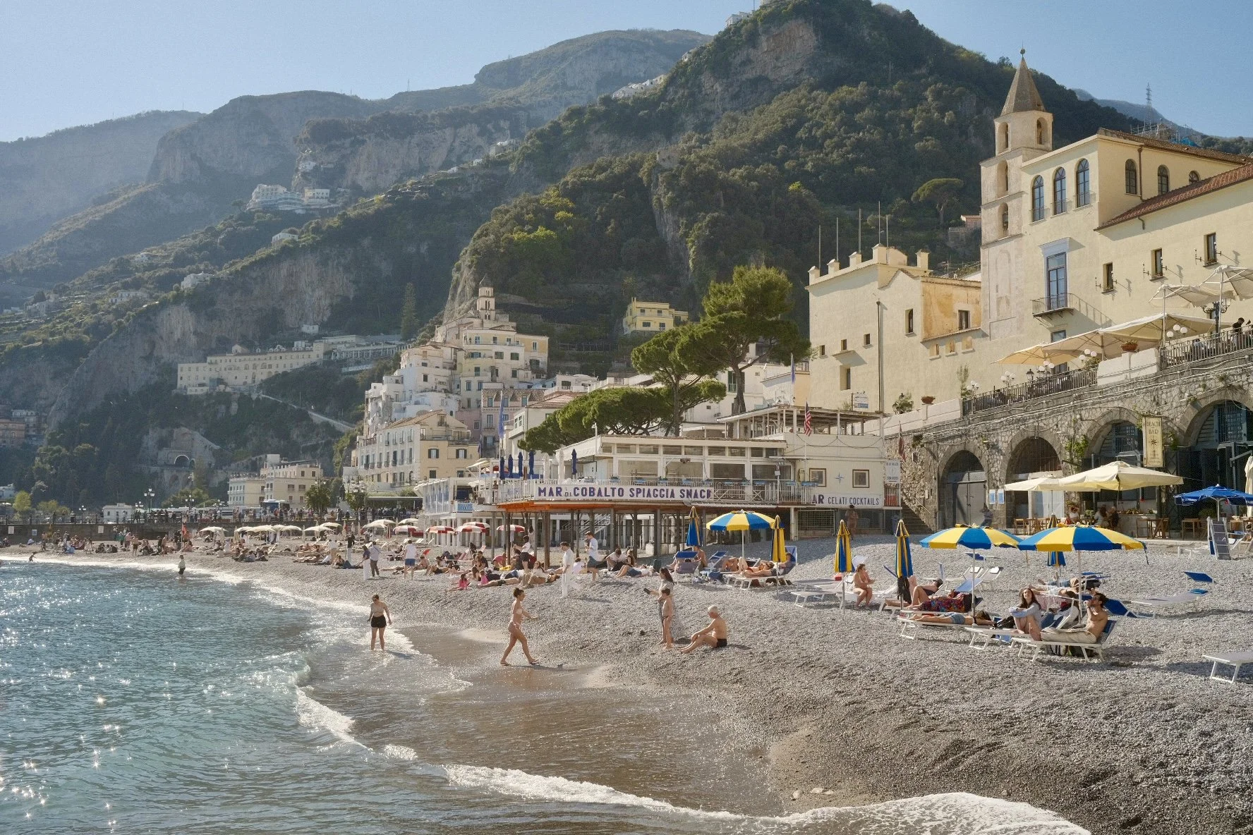 Beach scene with people sunbathing, umbrellas, and children playing by the water. In the background, there are colorful buildings and hillside with greenery and cliffs, with a church and other structures.