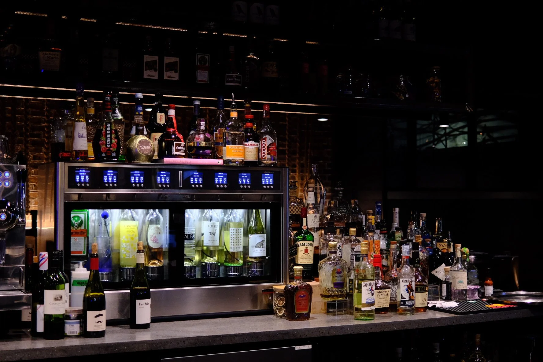 A bar with a variety of liquor bottles displayed on shelves, including whiskey, vodka, and other spirits, with a sleek refrigerated display case holding more bottles and bar equipment behind the counter.