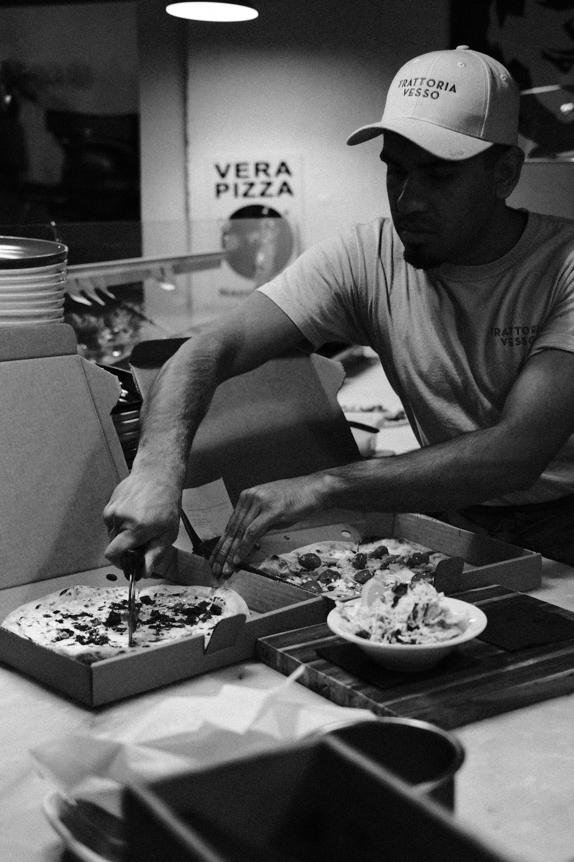 A man in a baseball cap and t-shirt prepares pizza in a restaurant kitchen. There are pizzas in boxes and plates on the counter, with a sign reading "Vera Pizza" in the background.