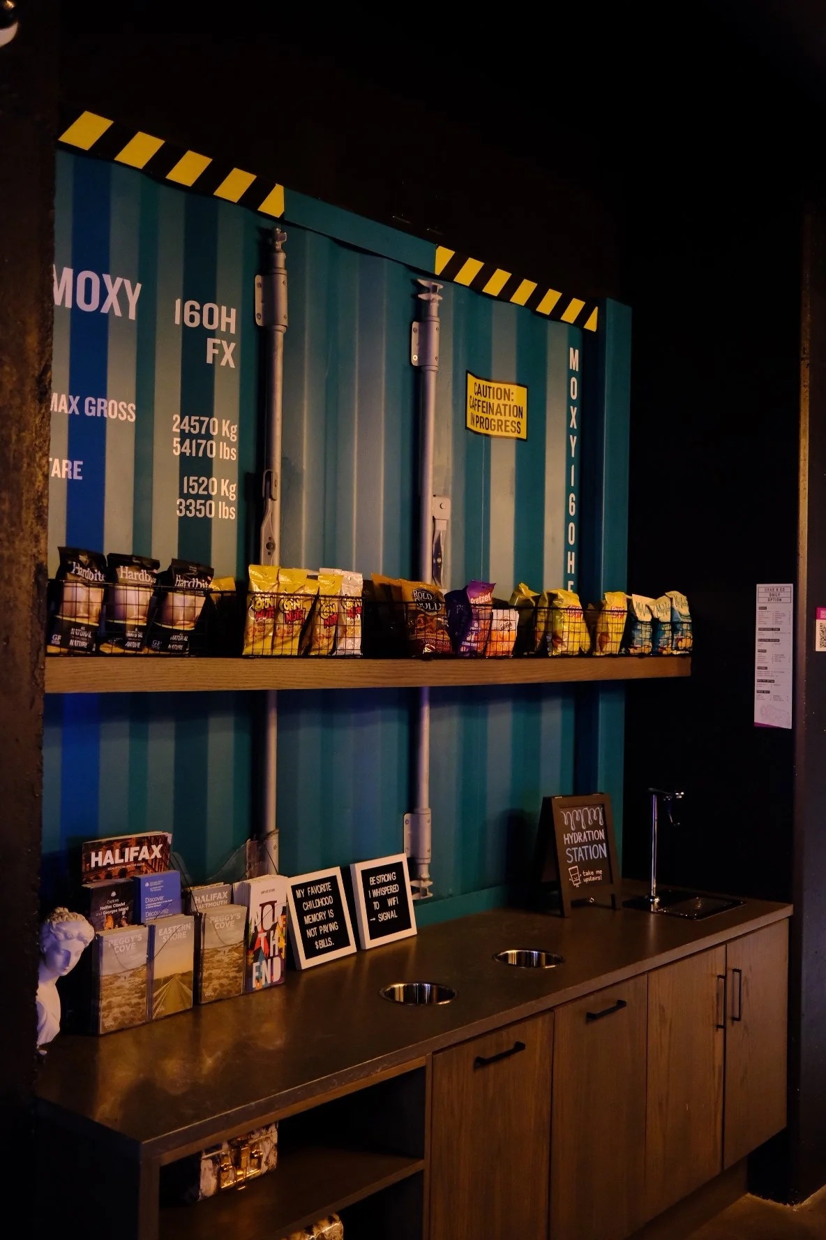 A display area with a blue shipping container wall, snack bags on a shelf, pamphlets, and signs including a hydration station sign.