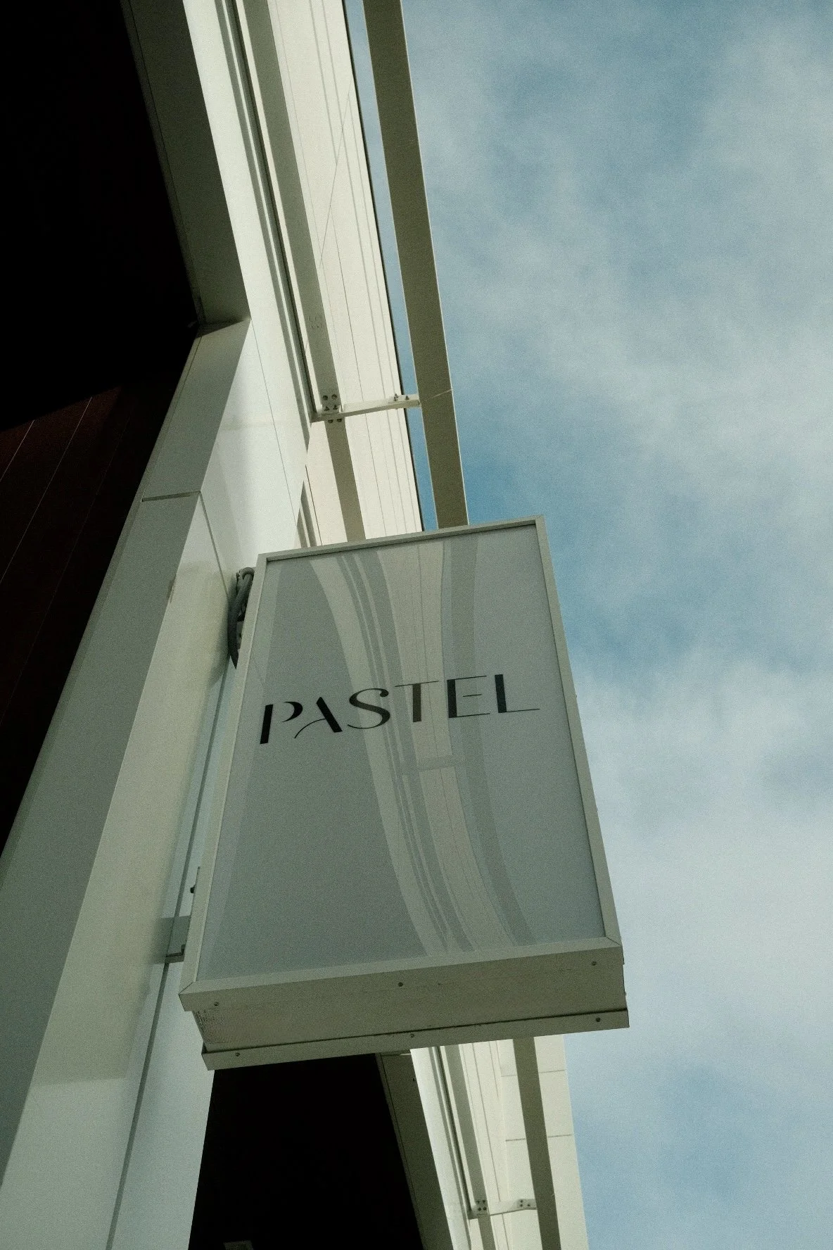 White sign with the word "PASTEL" attached to a building balcony with a view of the cloudy sky.