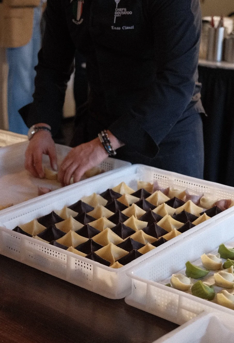 Person in black apron preparing colorful tortellini pasta on a table with trays of arranged finished pasta.