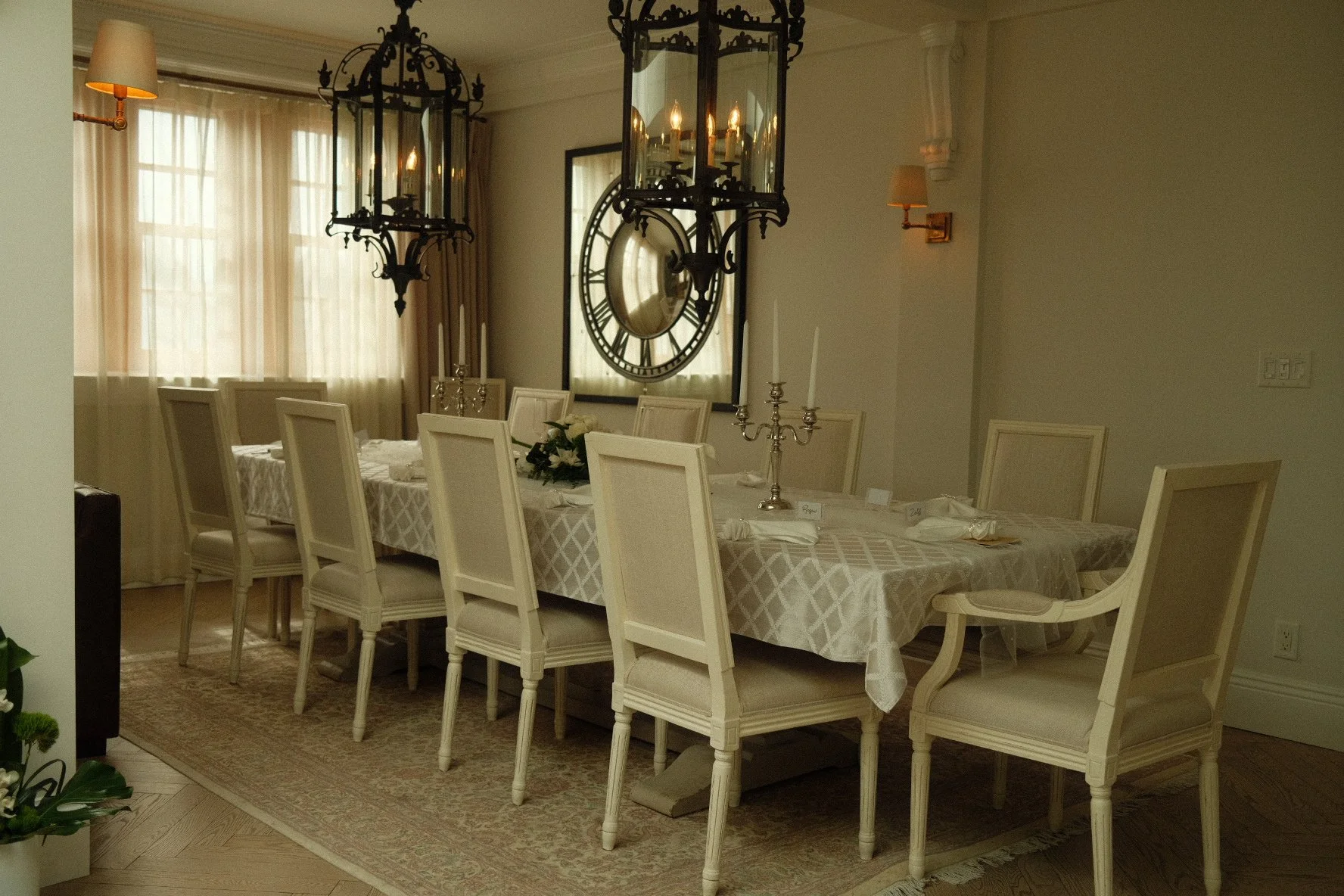 Elegant dining room with a long table covered in a patterned tablecloth, surrounded by white chairs with upholstered seats. The table is set with folded napkins and candelabras. The room features large windows with sheer curtains, ornate chandeliers 