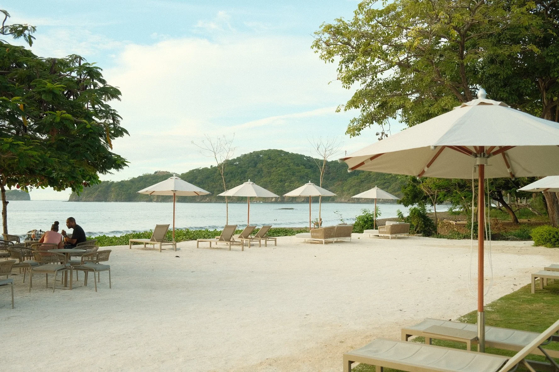 A beach scene with lounge chairs and umbrellas facing the ocean, some people sitting at tables, trees on either side, and lush green hills in the background under a partly cloudy sky.