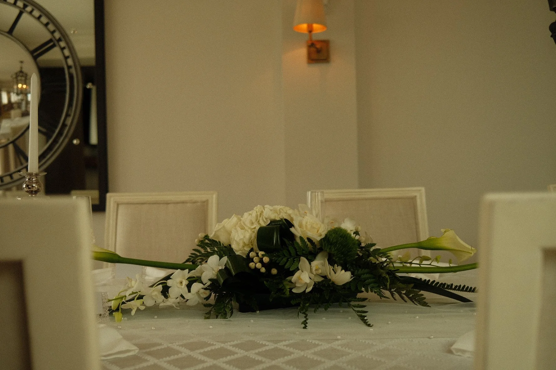 Elegant dining table centerpiece with white flowers and greenery, set in a well-lit room with beige chairs, a wall clock, and a wall-mounted light in the background.