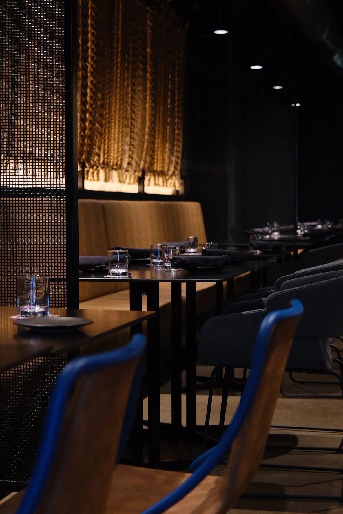A dimly lit restaurant interior with black tables, glasses of water, black napkins, and wooden chairs. Gold curtains cover the windows, and there is a decorative black partition.