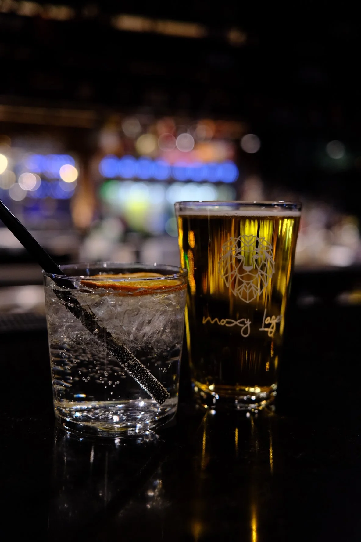 Two glasses filled with drinks on a dark bar counter, one with ice and a slice of lemon, the other with a lion logo and text in the background with blurry colorful lights.