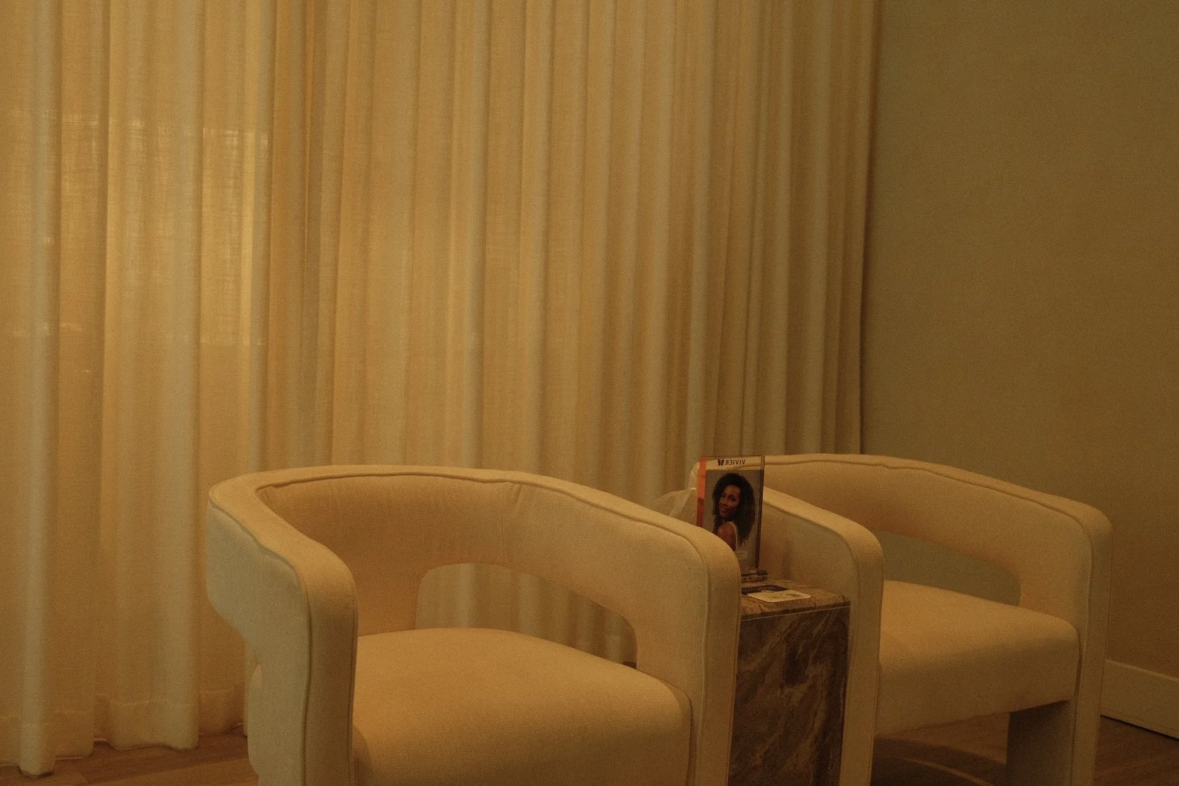 Two cream-colored armchairs with curved arms in front of beige curtains. A small marble table between them holds magazines and a pamphlet with a woman's photo.