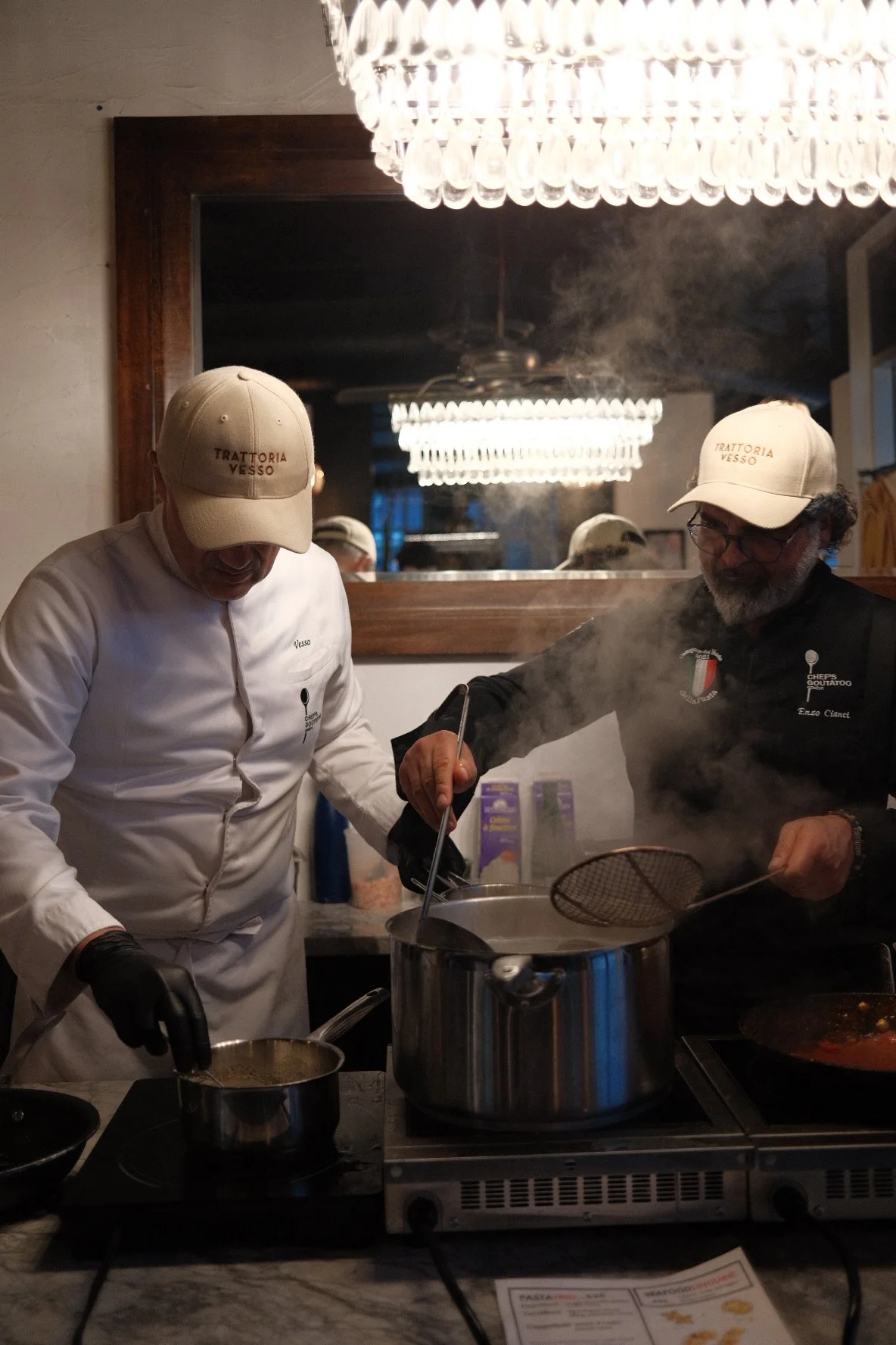 Two chefs cooking in a restaurant kitchen, one wearing a white coat and the other wearing glasses and a black jacket. Both are wearing beige hats with 'Trattoria Vesso' written on them. They are stirring pots on an induction stove, with steam rising,
