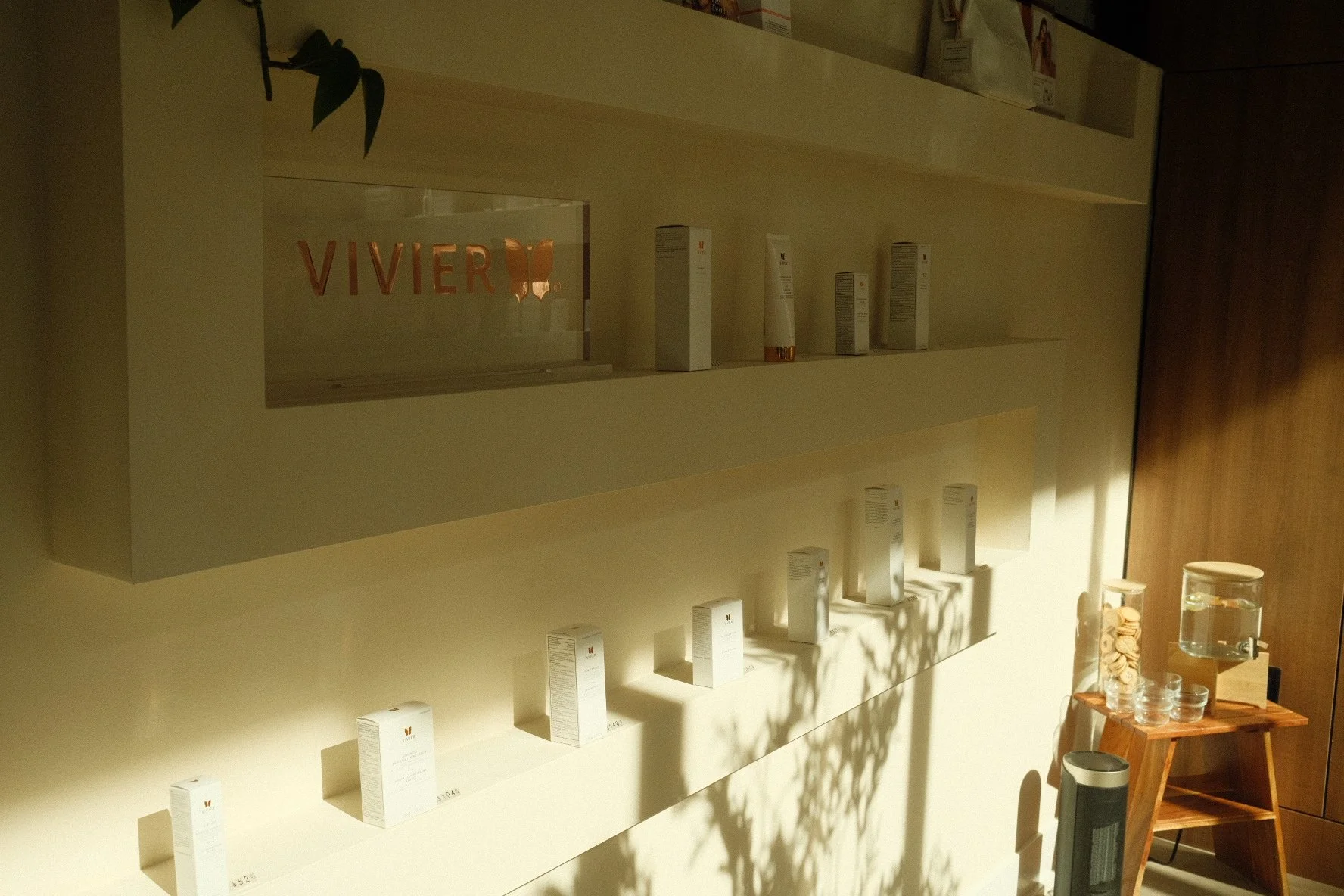 Display shelves with skincare or cosmetic products in a store, with a sign that reads 'VIVIER' and a butterfly logo, illuminated by natural light.