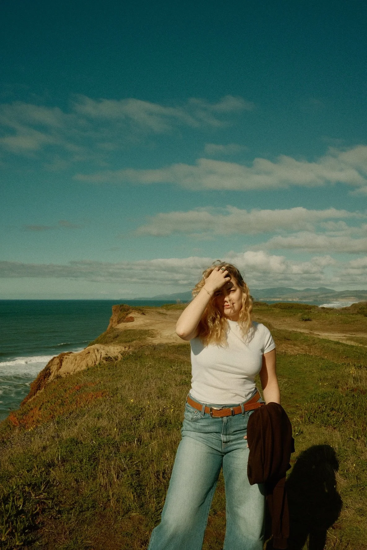 A woman with curly blonde hair standing on a grassy hillside near the ocean, holding her head with one hand, wearing a white T-shirt and light blue jeans, with a brown backpack hanging over her arm, scenic coastal landscape background with blue sky a