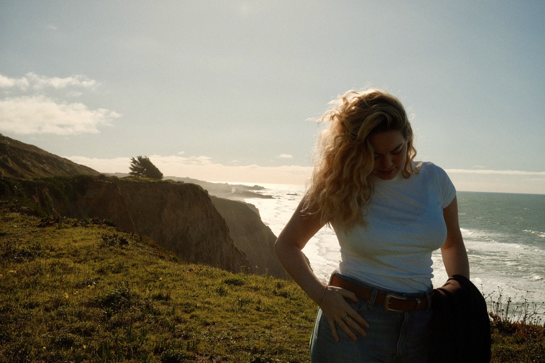 A woman with blonde curly hair looking down, standing on a grassy cliffside overlooking the ocean with waves and a distant shoreline, under a partly cloudy sky.