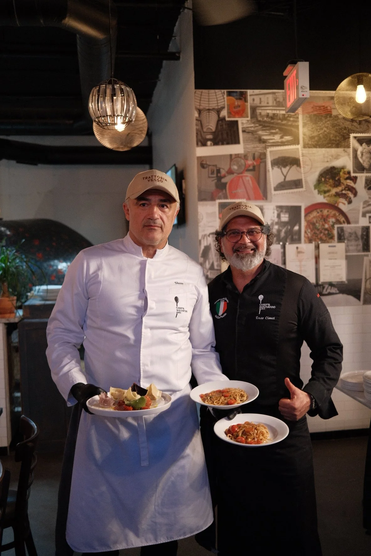 Two male chefs in a restaurant holding plates of pasta and dessert, smiling and dressed in chef uniforms with hats.