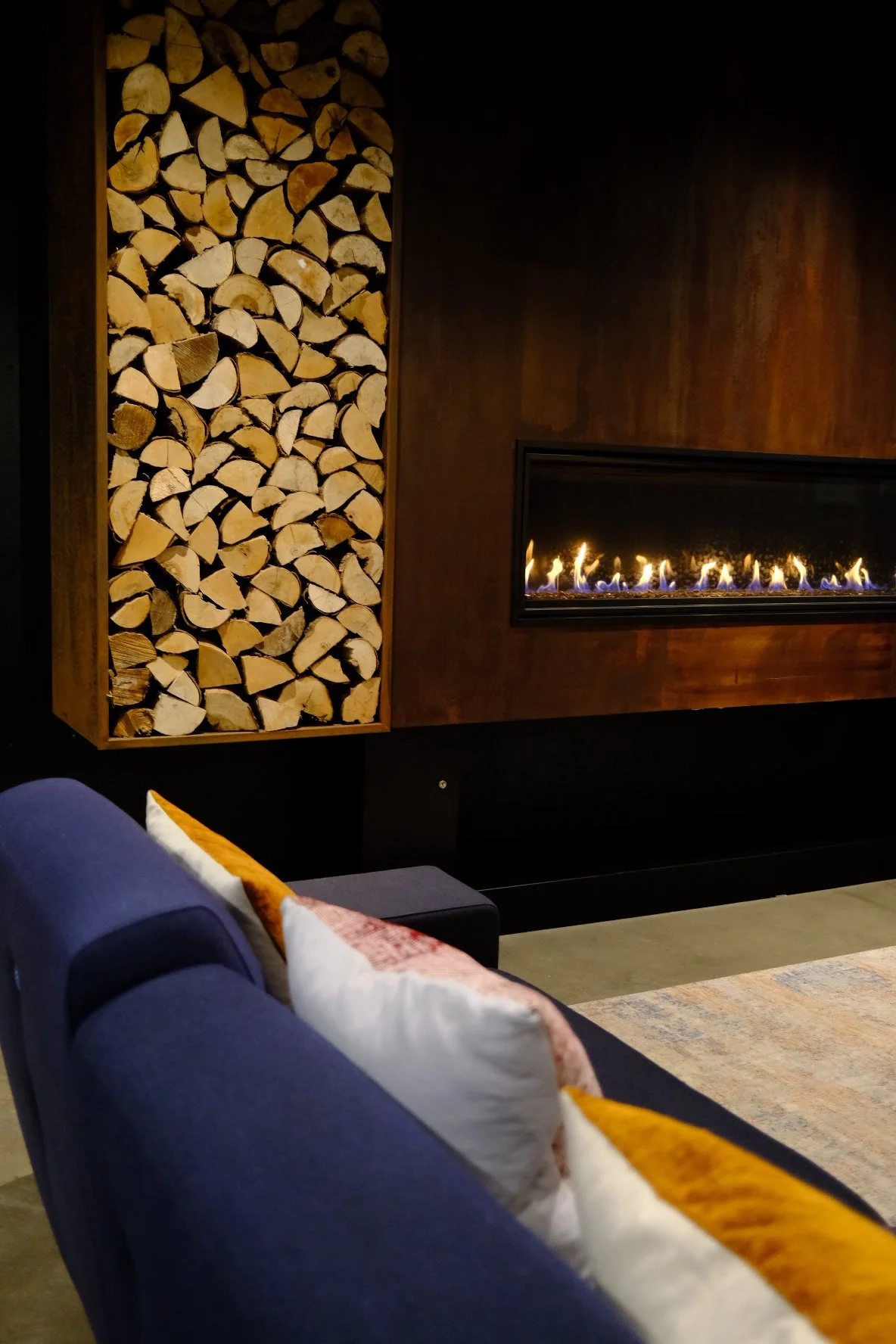 A cozy living room with stacked firewood in a built-in wooden storage and a modern rectangular fireplace with visible flames on a dark wall.