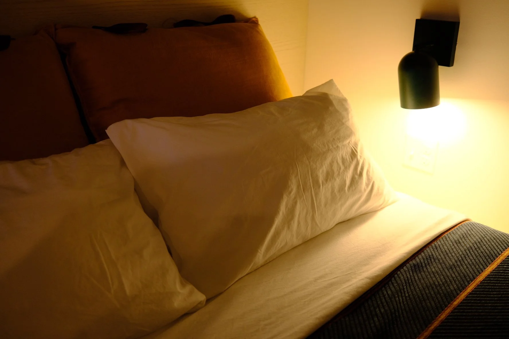 A neatly made bed with beige pillows and a beige bedspread, illuminated by a black wall-mounted reading lamp in a cozy room.