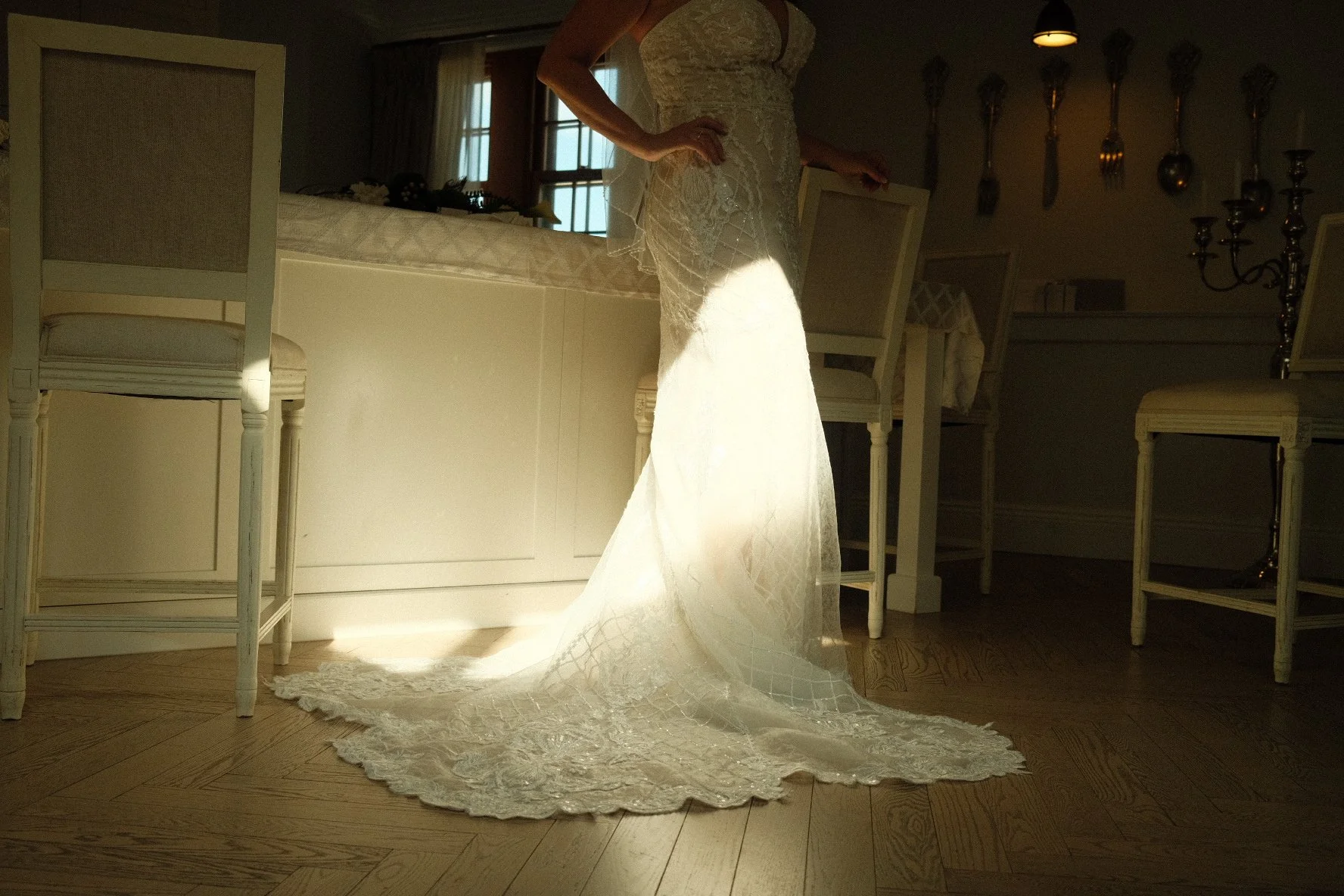 A white wedding dress with a long train draped on a chair in a dimly lit room with white chairs and a chandelier, with sunlight coming through a window.