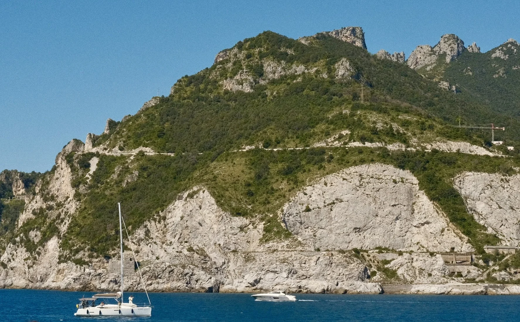 A sailboat and a motorboat on the water in front of a large rocky and green hillside under a clear blue sky.