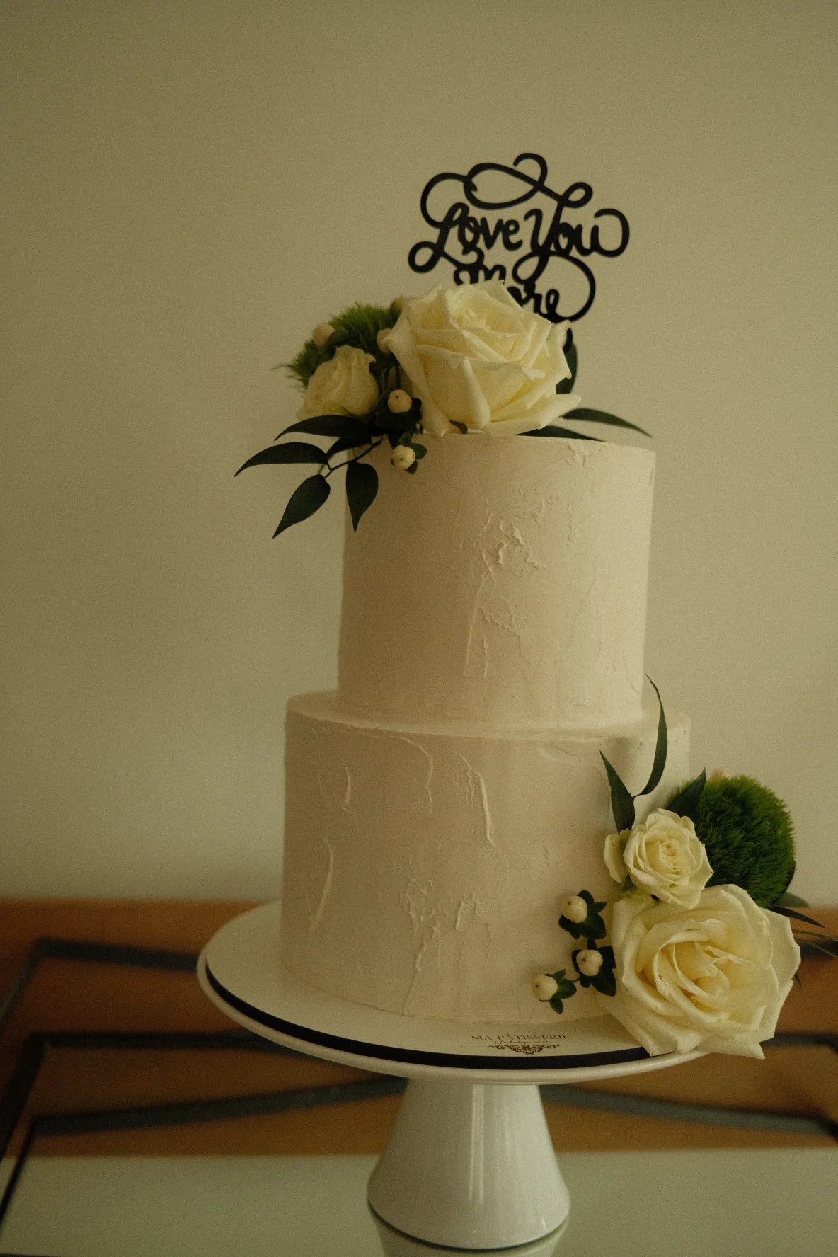 A two-tiered white wedding cake decorated with white roses, greenery, and a black topper that says "Love You More," placed on a white cake stand.