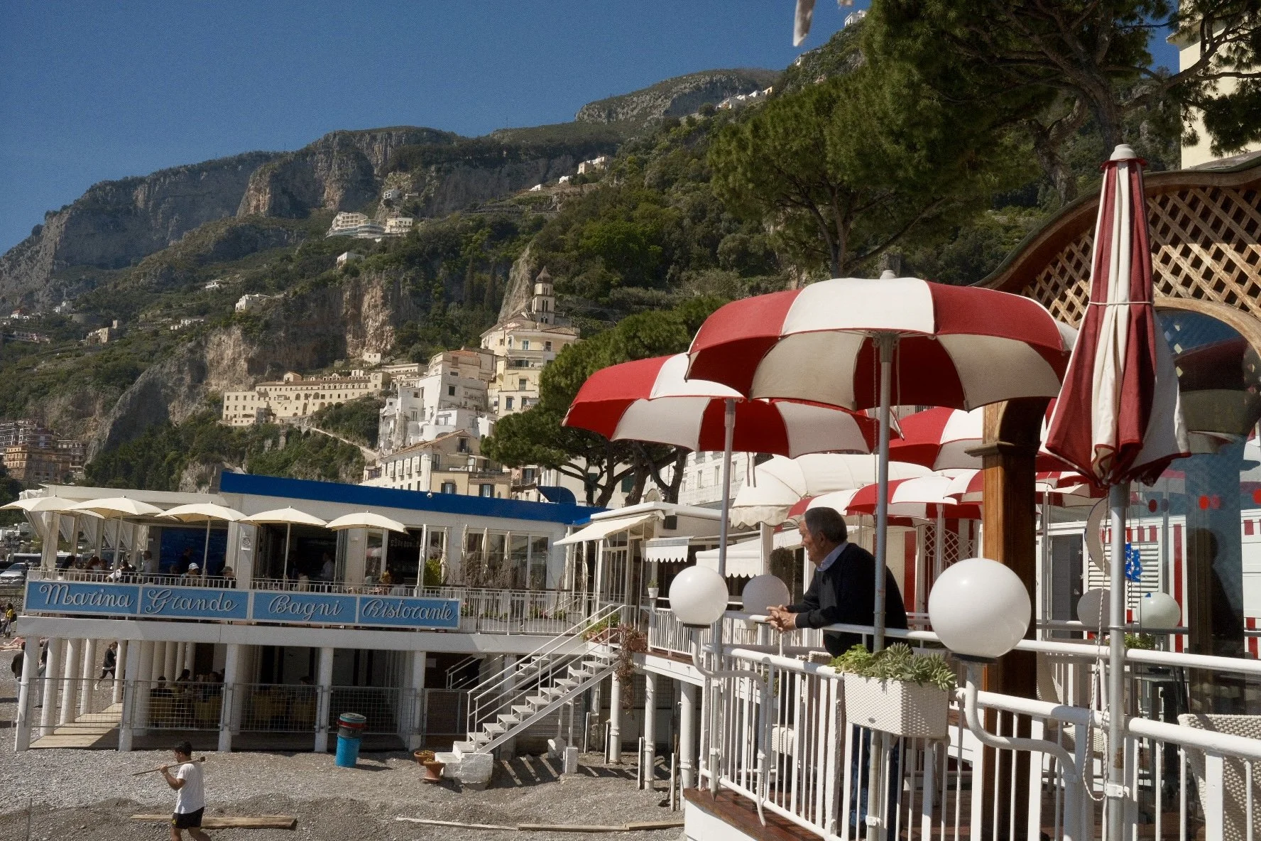 Beachside outdoor restaurant with red and white umbrellas, white railings, and stairs leading to a lower level. Hillside with white buildings and greenery in the background, under a clear blue sky.