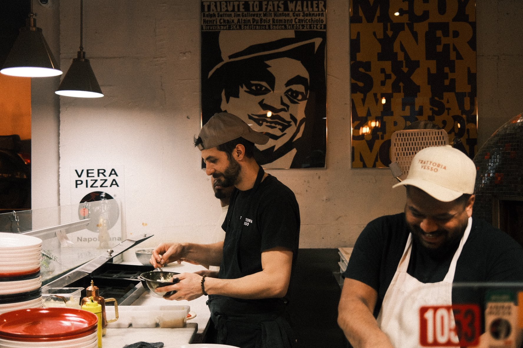 Two men working behind the counter in a restaurant or pizzeria, preparing food. One in a black cap and the other in a white cap with a smile. Posters and artistic images adorn the wall behind them.