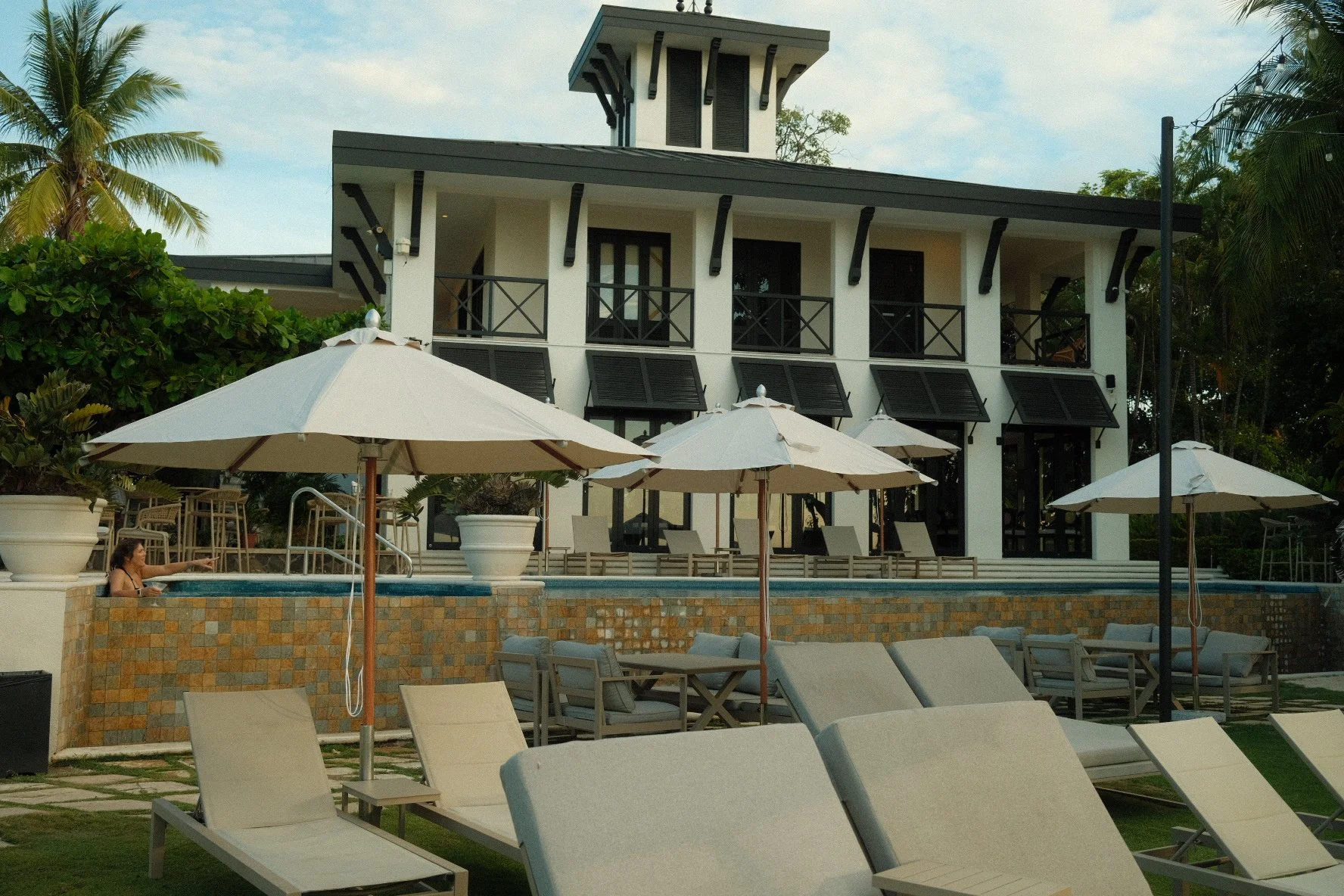 A resort pool area with white lounge chairs and umbrellas in the foreground, a person relaxing in the pool, and a modern white building with black accents, balconies, and large windows in the background, surrounded by green trees and tropical plants.