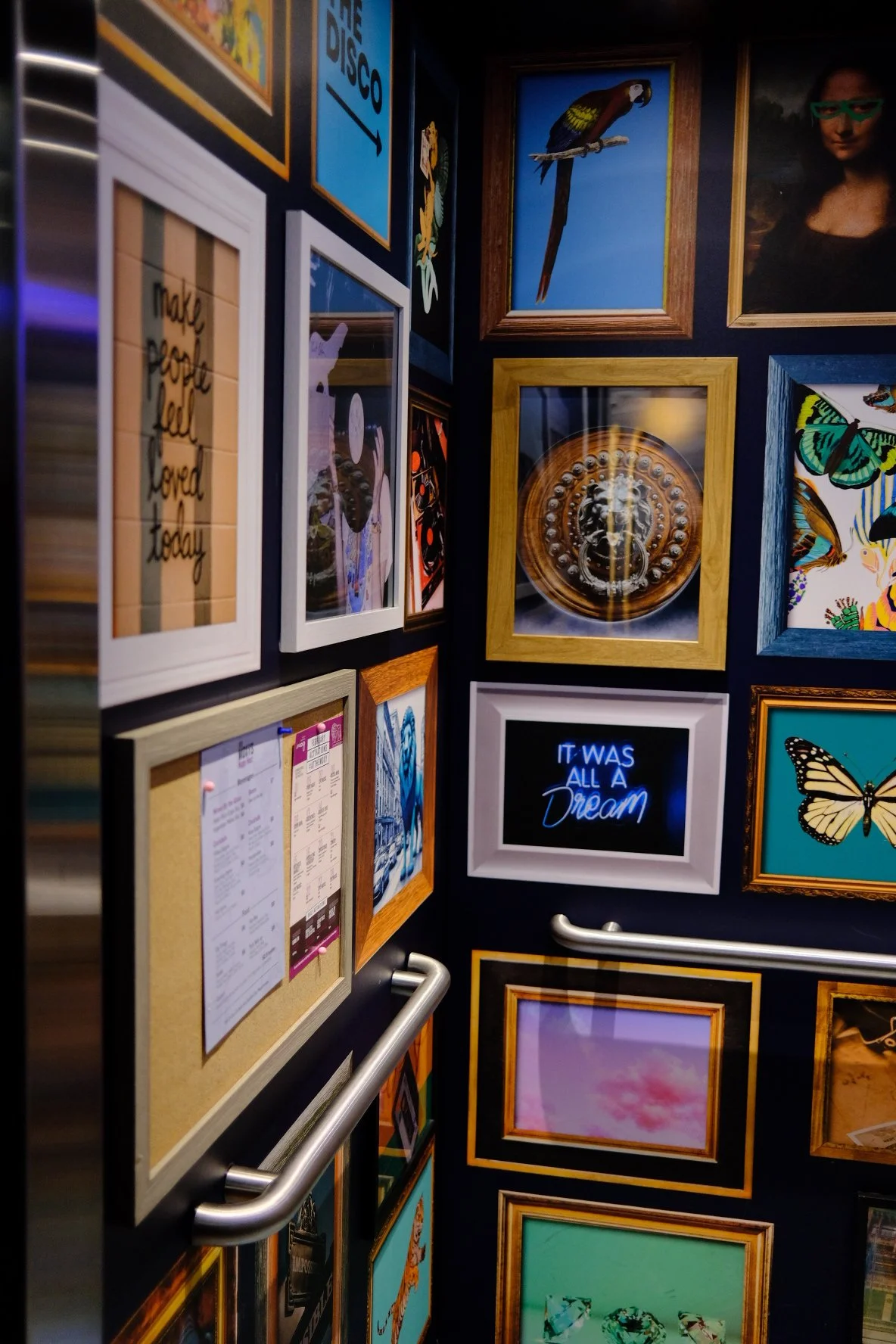 A corner of a gallery wall with various framed artworks and photographs, including a parrot, a famous painting, butterflies, and neon signs, on a dark background.