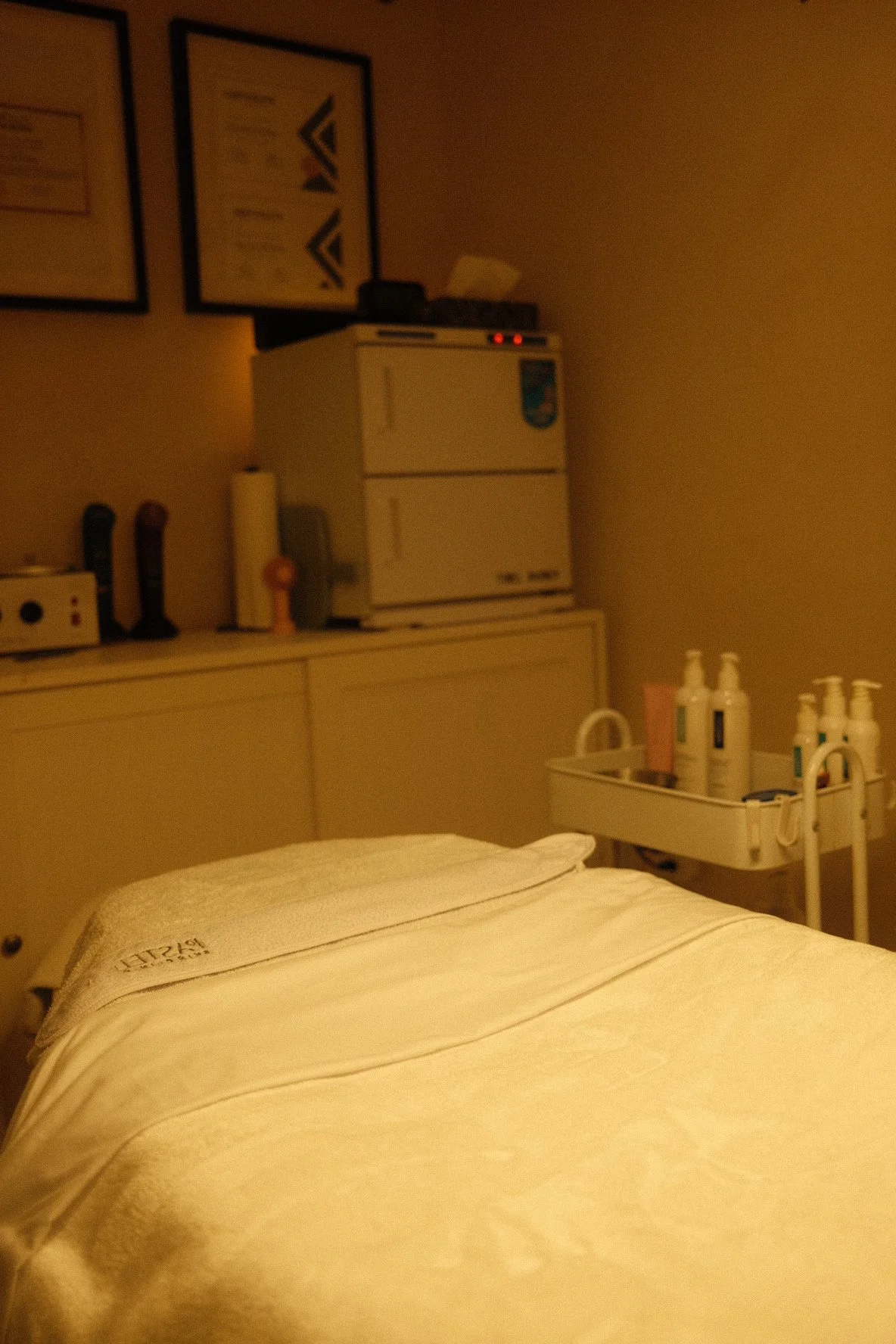 A hospital room with a bed, medical supplies, and a medical cart. The image shows a bed with a white blanket in the foreground, a tray with lotion bottles, and a cabinet with medical equipment in the background.