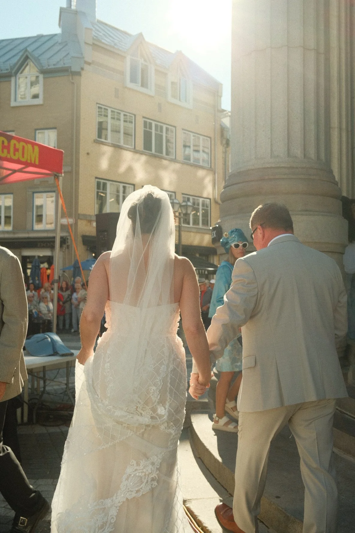 A bride and groom holding hands during their wedding ceremony outdoors with a crowd and buildings in the background.