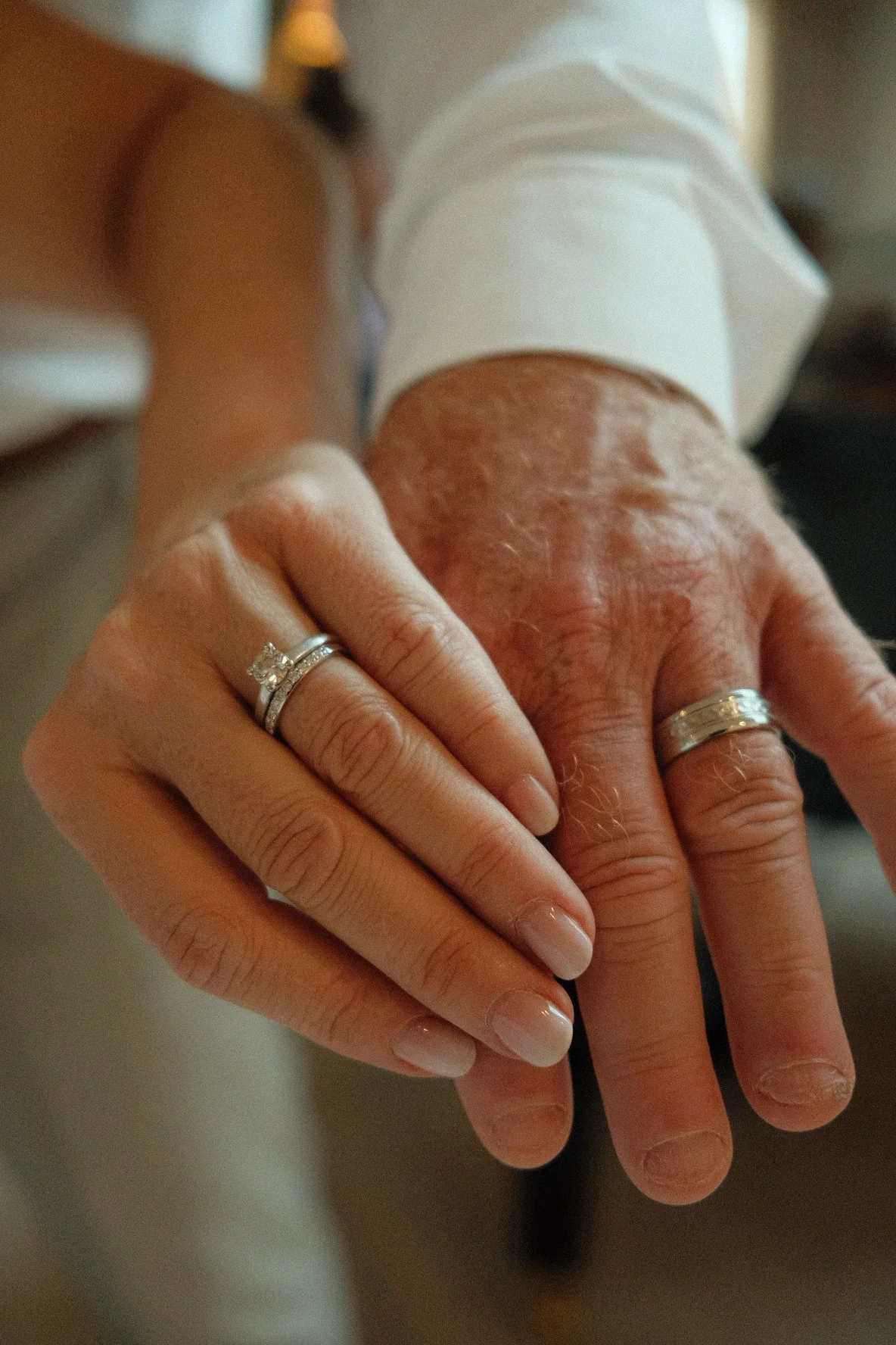 A close-up of a person's hand with wedding and engagement rings, resting on an older person's hand with visible skin texture.