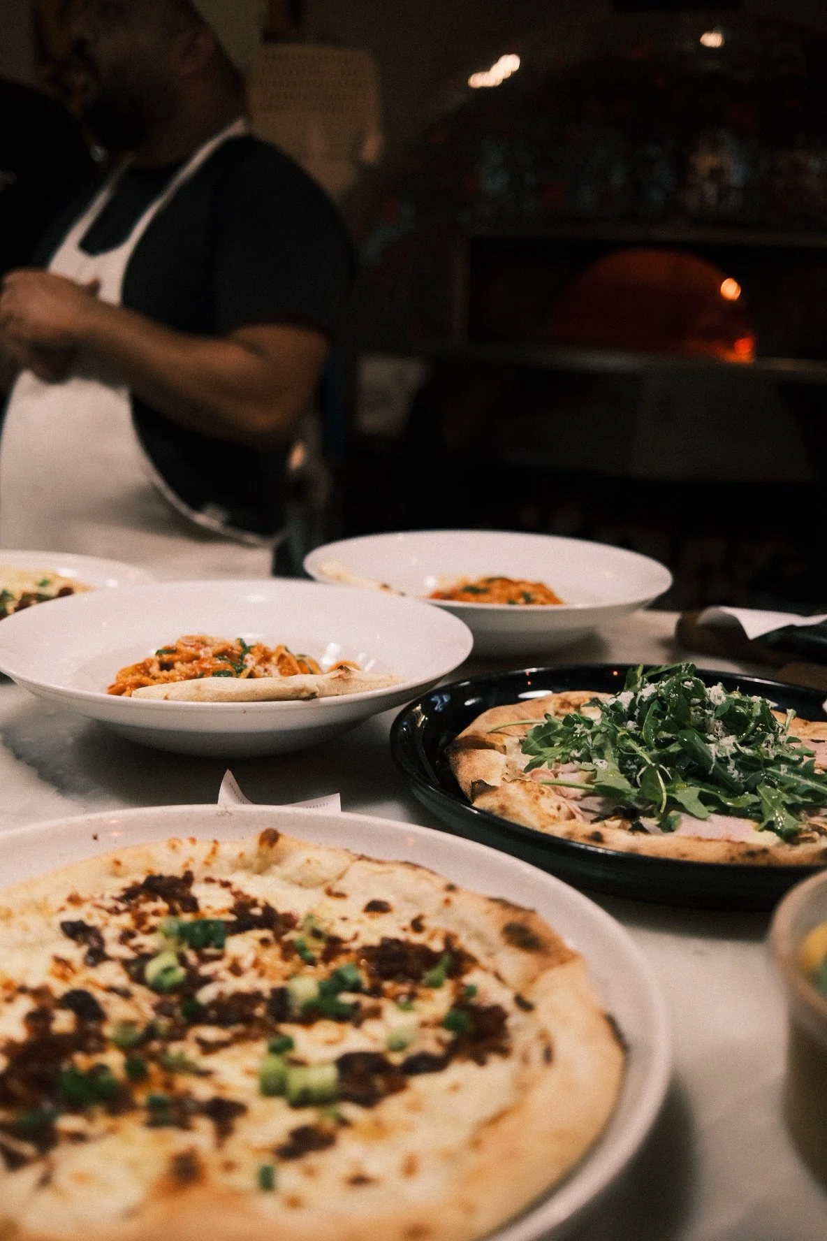 Multiple plates of pizza and bowls of pasta on a table in a restaurant with a chef working in the background.