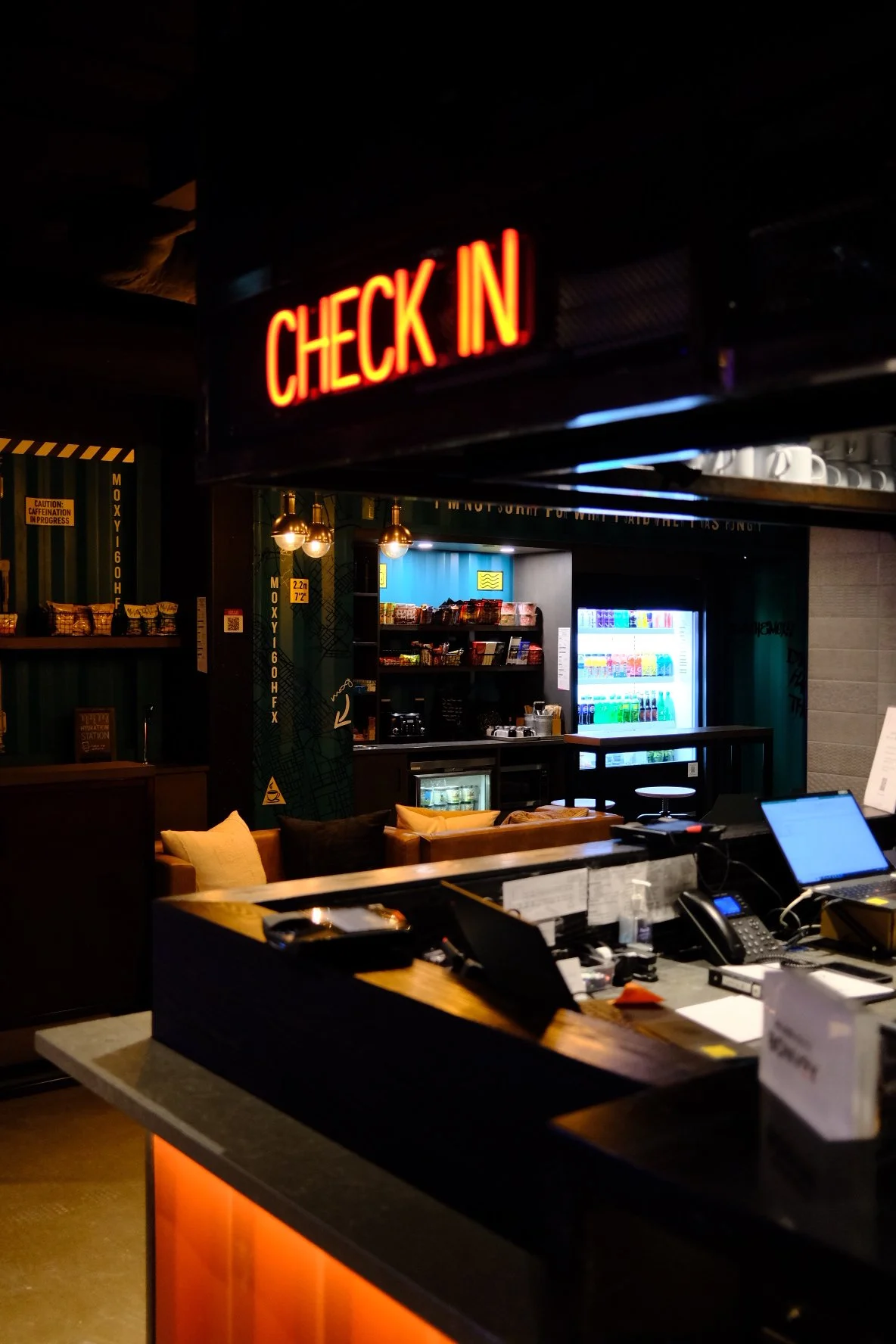 A check-in area in a hotel or restaurant with a red neon sign that says 'CHECK IN'. The background shows a vending machine with beverages, a small kitchenette, and comfortable seating with pillows.