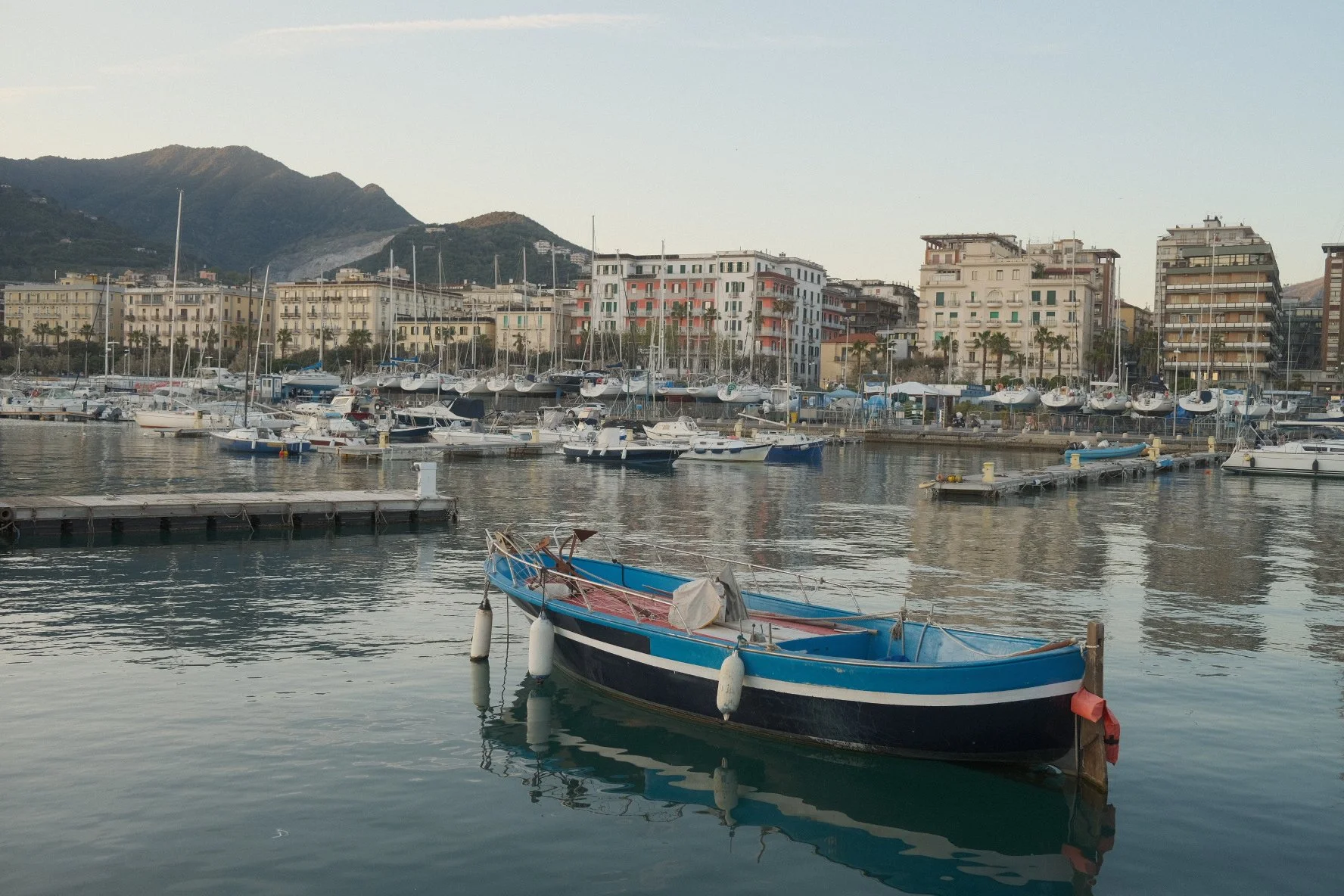A small harbor with sailboats and yachts docked, with residential buildings and green hills in the background.
