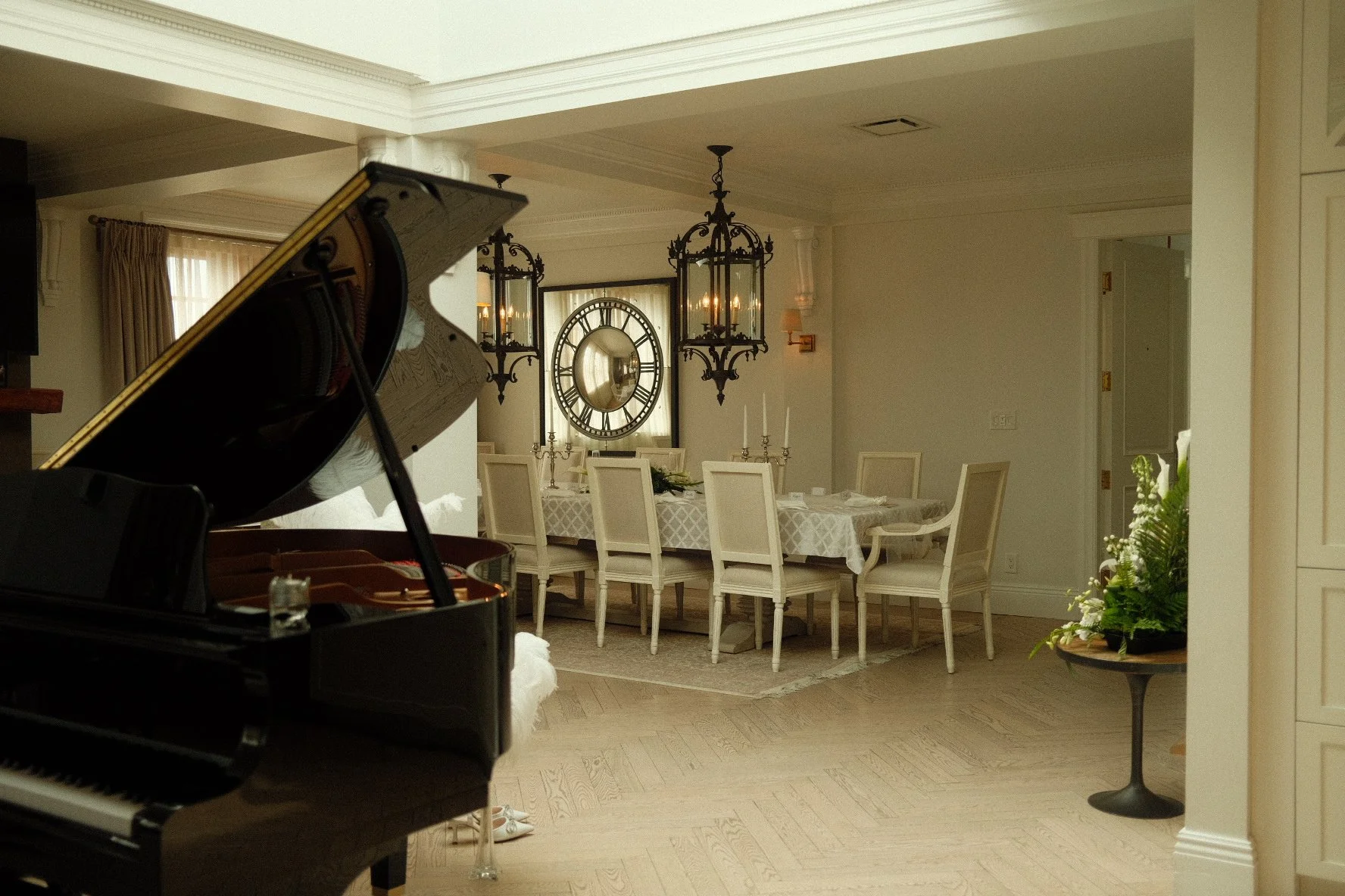 A dining room with a large clock on the wall, a table set for a meal with white chairs, a chandelier, and a piano in the foreground.