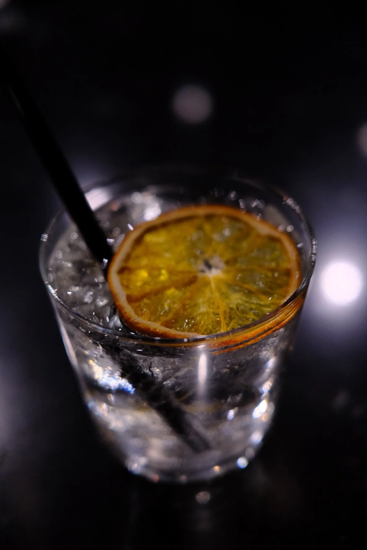 A close-up of a glass filled with a clear beverage, ice cubes, a slice of dried orange, and a black straw on a dark surface.