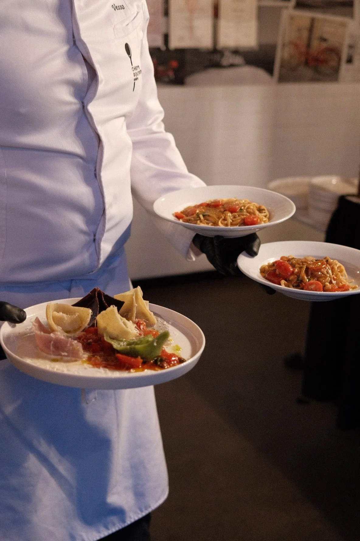 Person wearing a white chef's coat and black gloves holding plates of pasta and a cheese platter with various cheeses and prosciutto in a restaurant or kitchen setting.