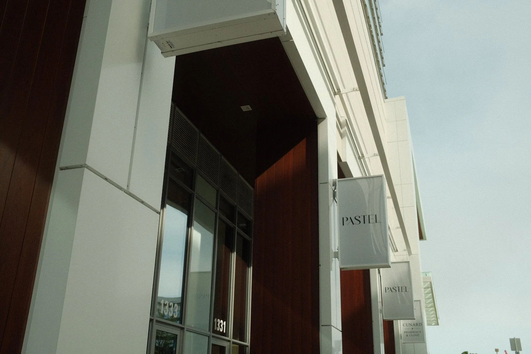 Exterior view of a modern building with white and wood-paneled walls, glass windows, and signs that read 'PASTEL' and 'CUNARD PHARMACY & CLINIC'.